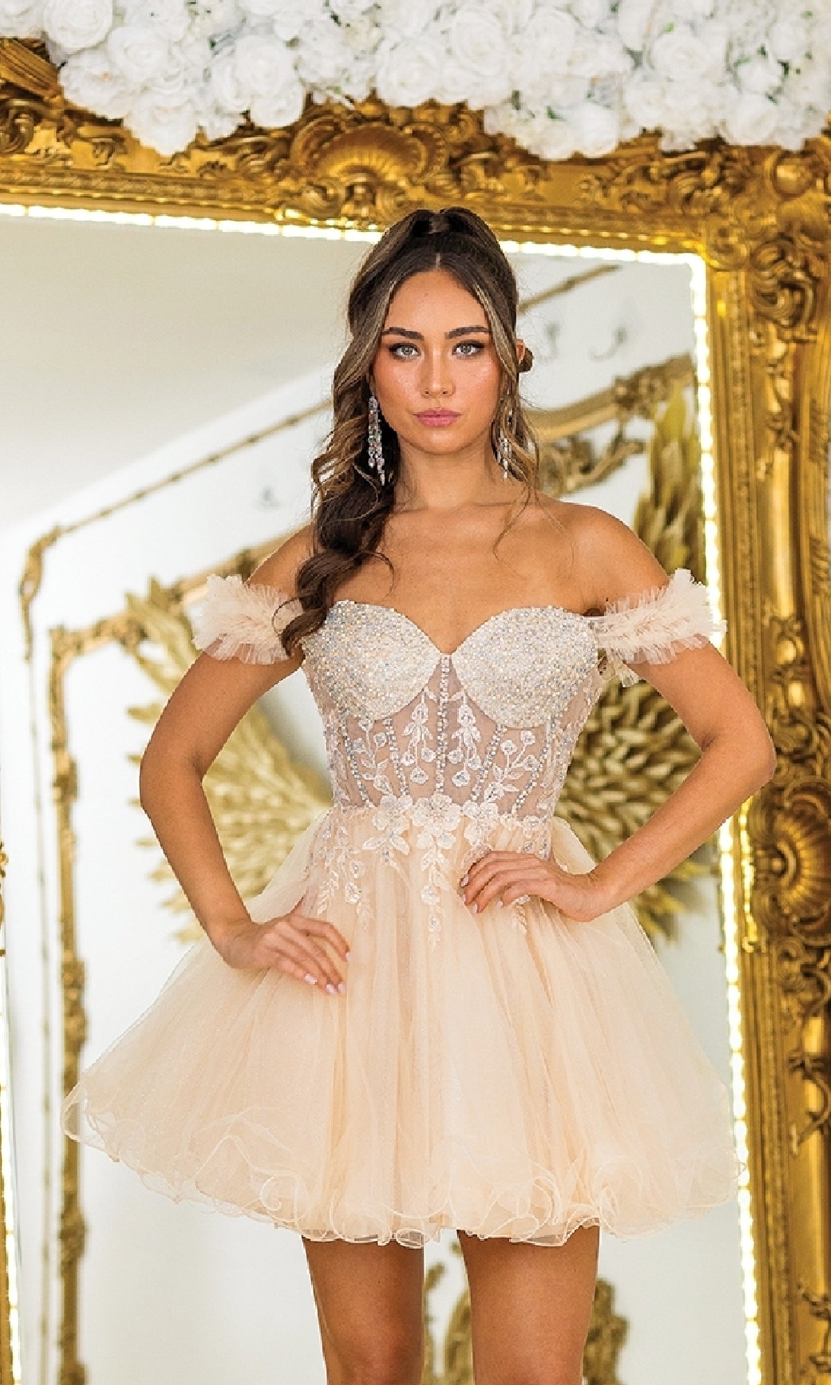 A woman stands before a gold ornate mirror, wearing the Dancing Queen 3406 Ruffle Short A-Line Prom Dress with an off-shoulder blush pink embellished bodice and tulle skirt. White roses are displayed above the mirror.