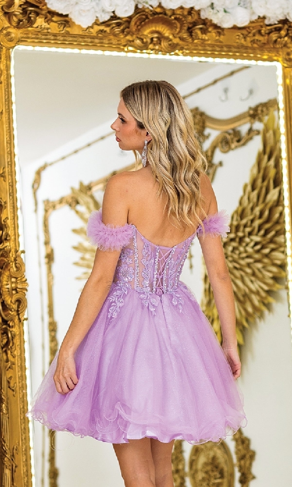 A woman in the Ruffle Short A-Line Prom Dress: Dancing Queen 3406 stands before an ornate gold-framed mirror with decorative wing details in the background.
