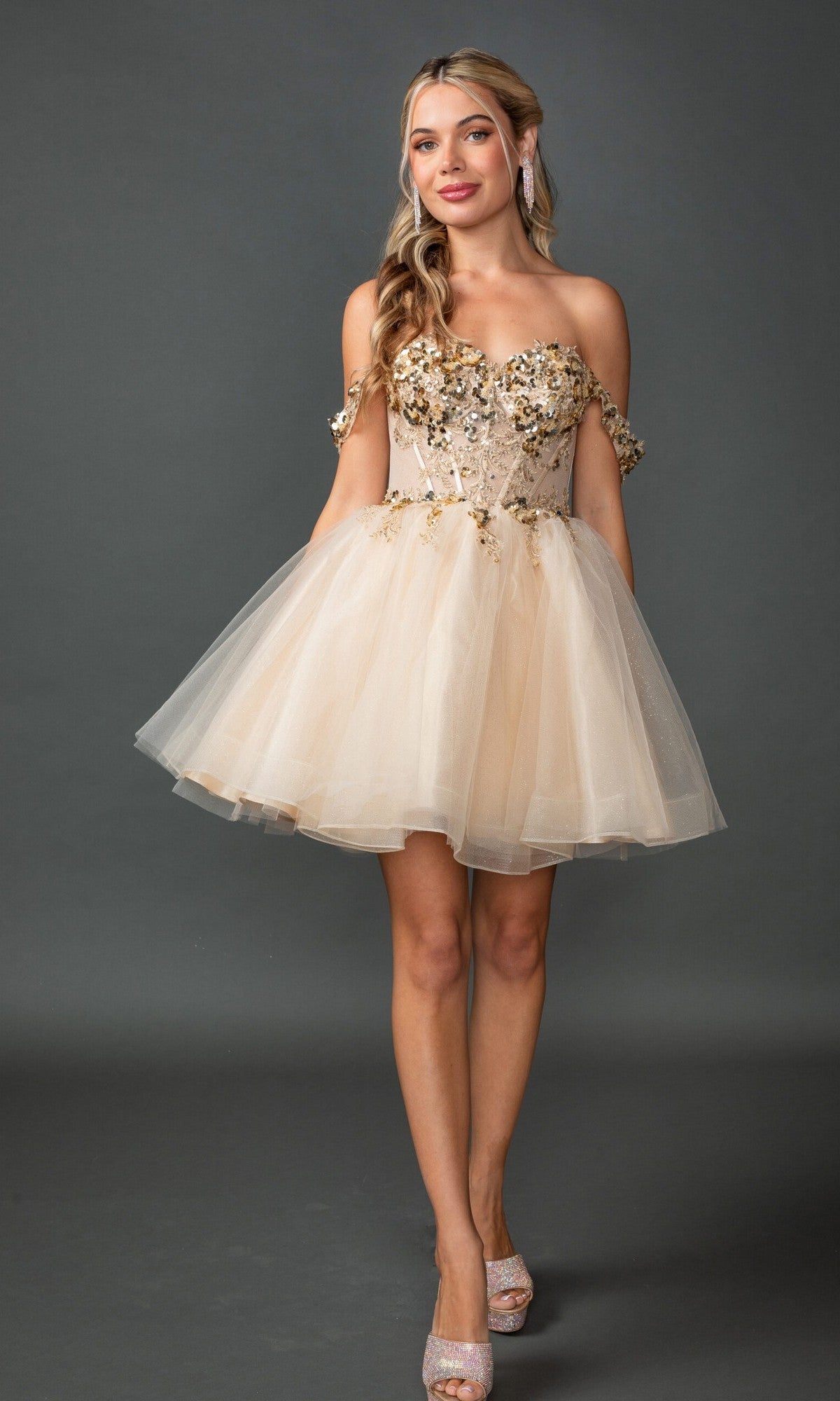 A woman is wearing the Off-Shoulder Glitter Hoco Dress: Dancing Queen 3408 in champagne, an elegant off-the-shoulder tulle dress with floral embellishments, standing against a gray background.