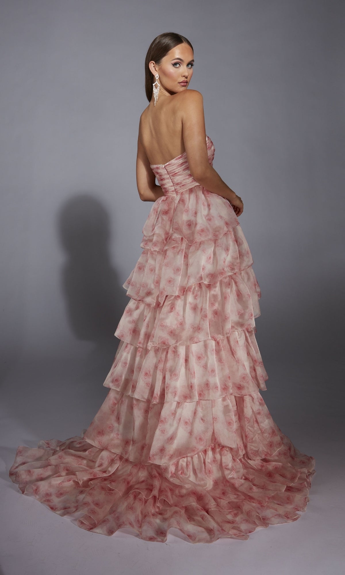 A woman stands against a gray background, wearing the Pink Floral Prom Ball Gown: Aerie Couture 340 with a strapless sweetheart ruched bodice, tiered pink floral layers, and a long train as she looks over her shoulder.