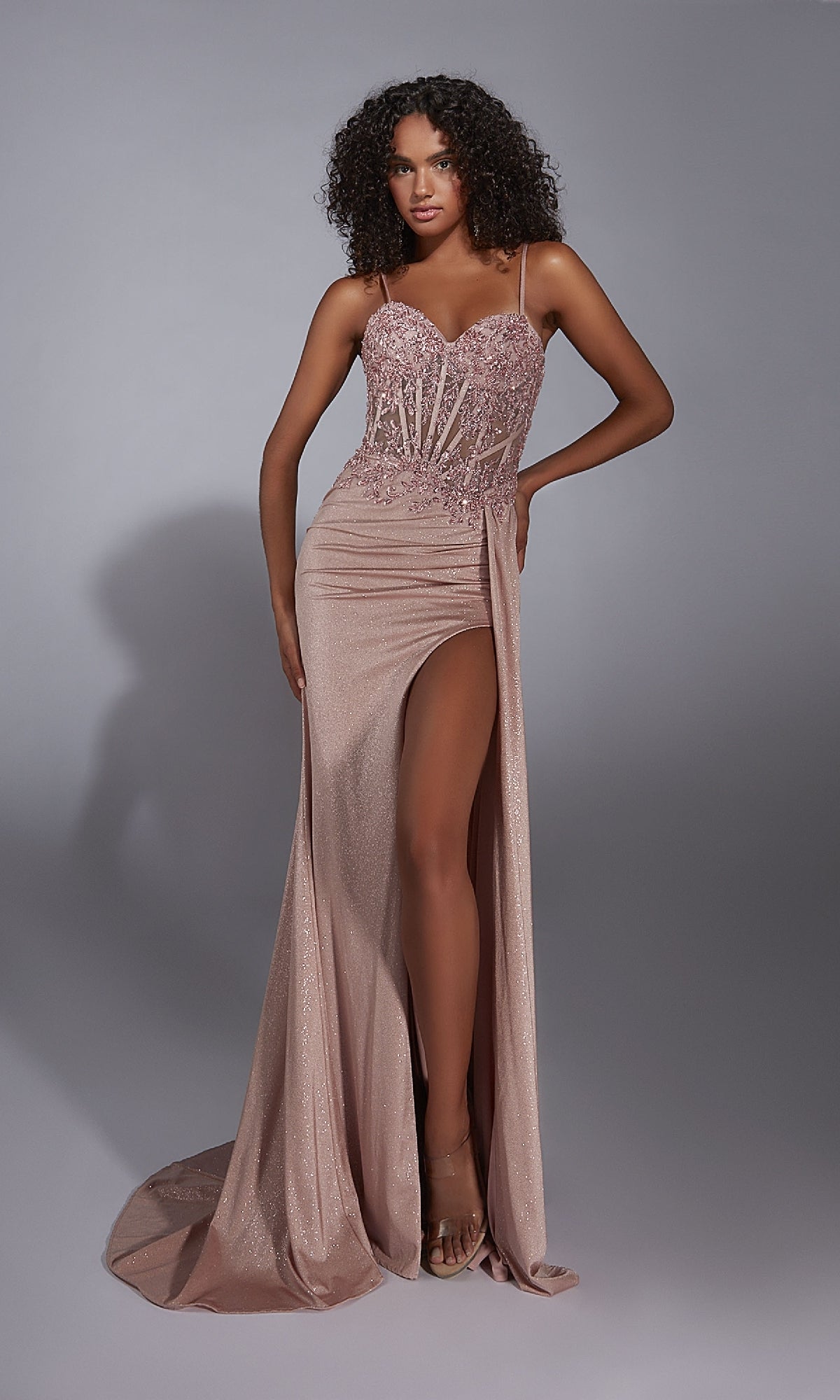 A woman poses in the Aerie Couture 341 long prom dress, featuring pink glitter fabric, a high slit, thin straps, and a beaded lace corset bodice, set against a plain gray background.
