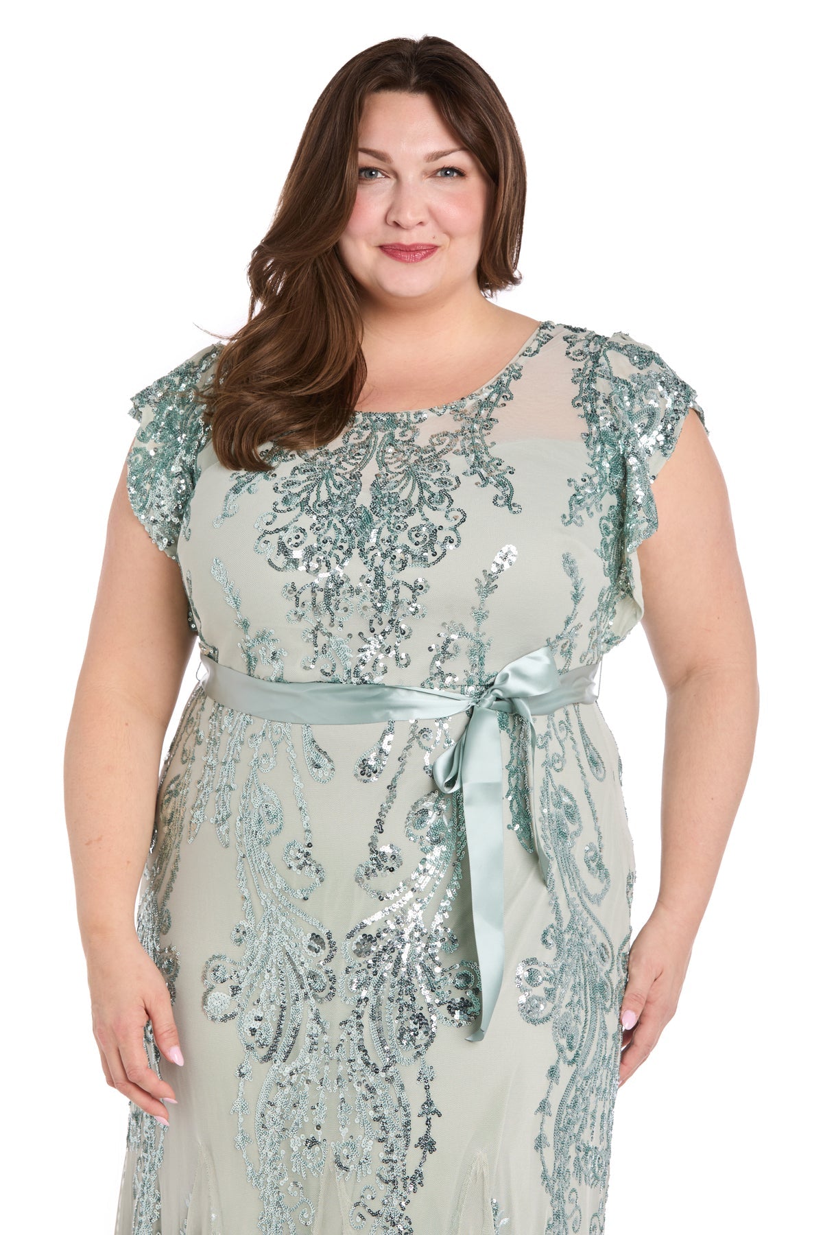 A woman stands smiling against a white background, wearing the Plus-Size Sage Green Formal Dress: R&M Richards 3465W, featuring sequin print, short sleeves, and a satin waist tie.