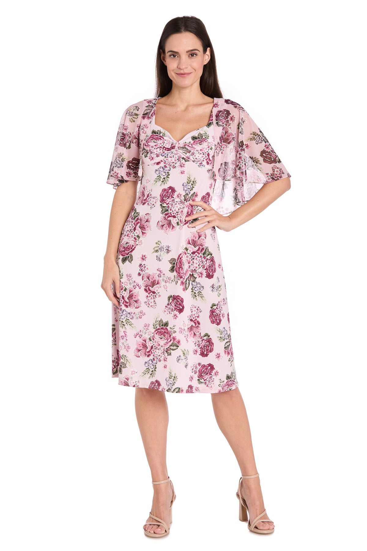 A woman wears the R&M Richards 3467P Petite Caped Pink Party Dress with light floral prints and a sheer, short-sleeved cover-up, paired with nude heels—an elegant semi-formal look against a white background.