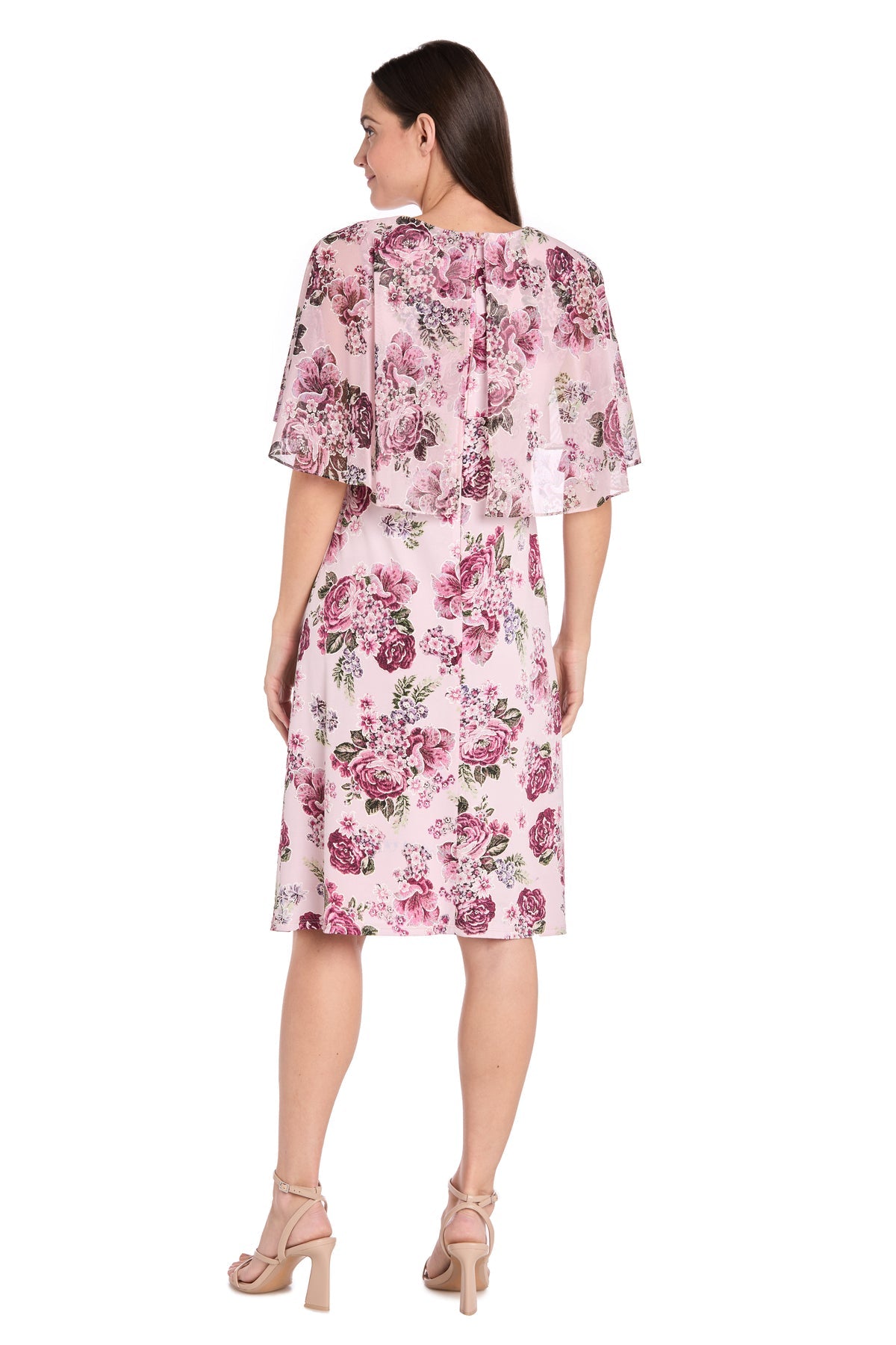 A woman, seen from behind, wears the R&M Richards 3467P Petite Caped Pink Party Dress with floral prints and short sleeves, paired with nude high-heeled sandals against a white background.