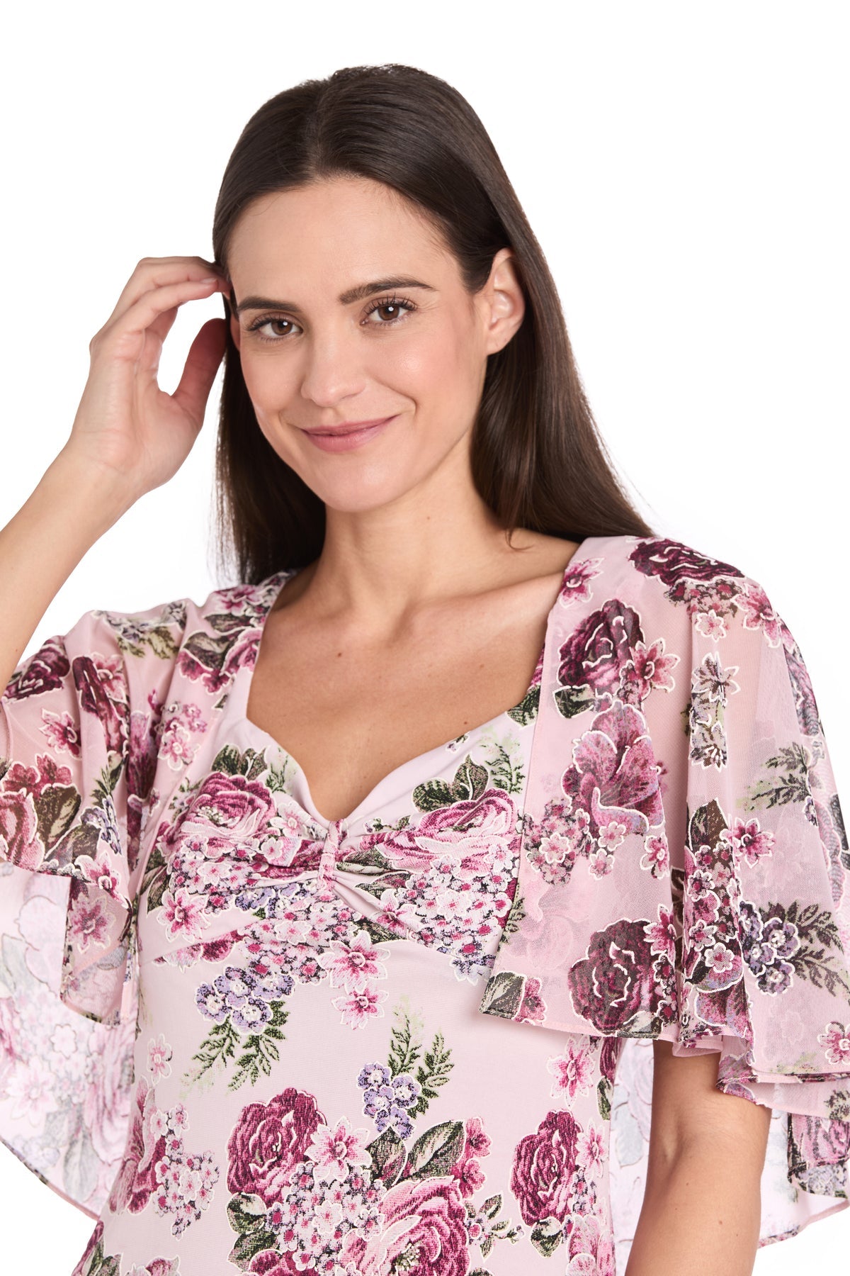 A woman with long dark hair wears the R&M Richards 3467P Petite Caped Pink Party Dress, featuring sheer, flowy sleeves, as she stands against a white background and touches her hair.