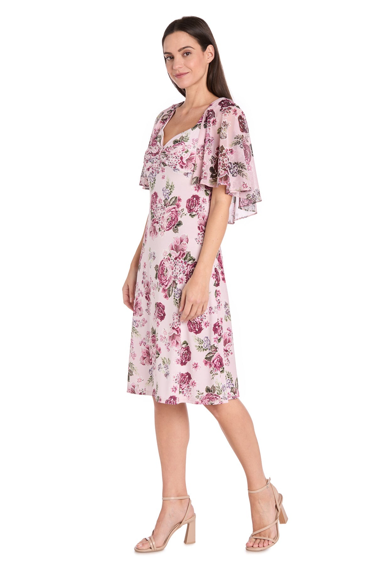 A woman poses in the Petite Caped Pink Party Dress: R&M Richards 3467P, a knee-length light pink floral print cocktail dress with flutter sleeves and nude heels, perfect for semi-formal occasions against a white background.