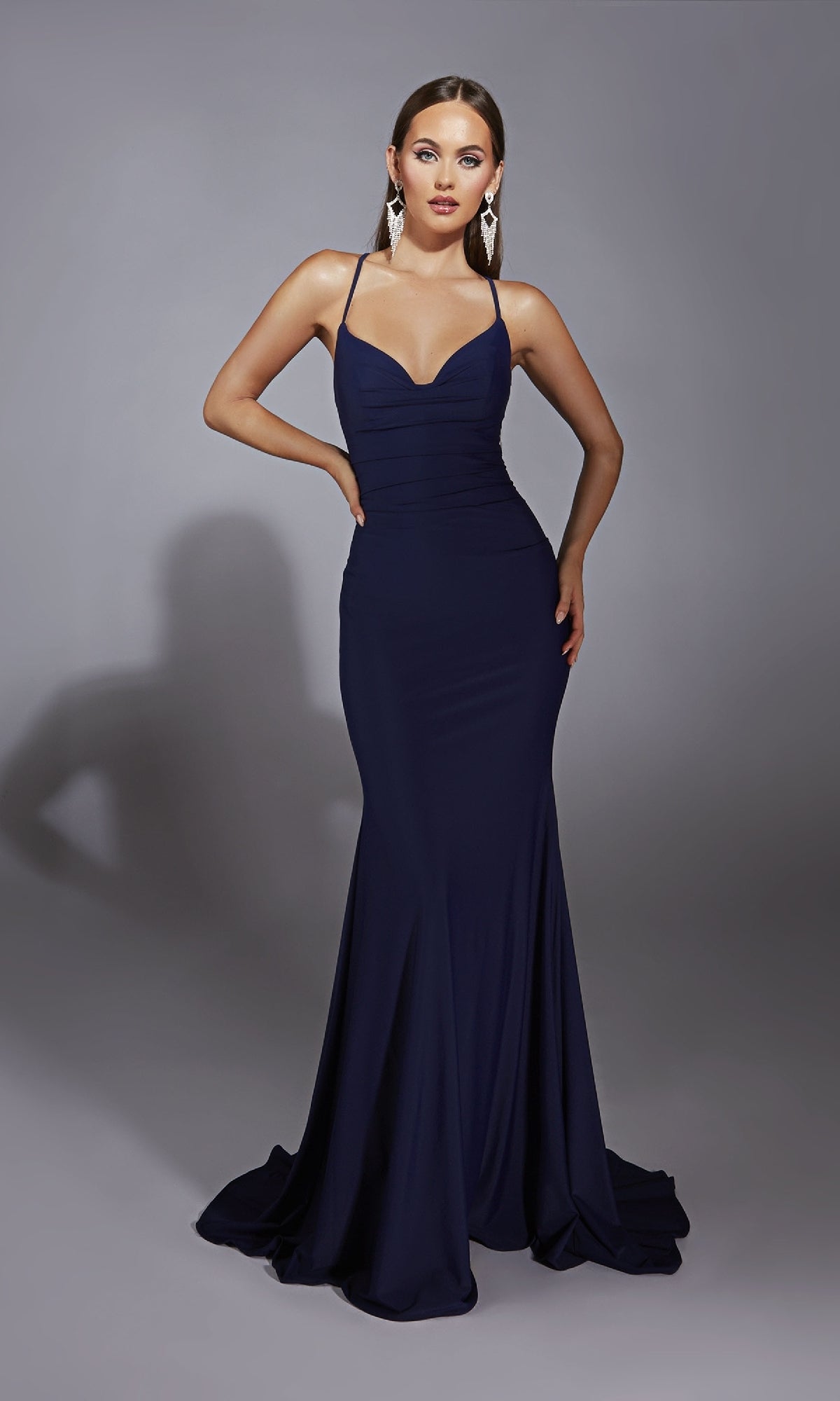 A woman poses in the Aerie Couture 346 Cowl-Neck Long Prom Dress, a fitted navy blue gown with spaghetti straps and an open back, styled with statement earrings against a gray background. This formal dress exudes sophistication.
