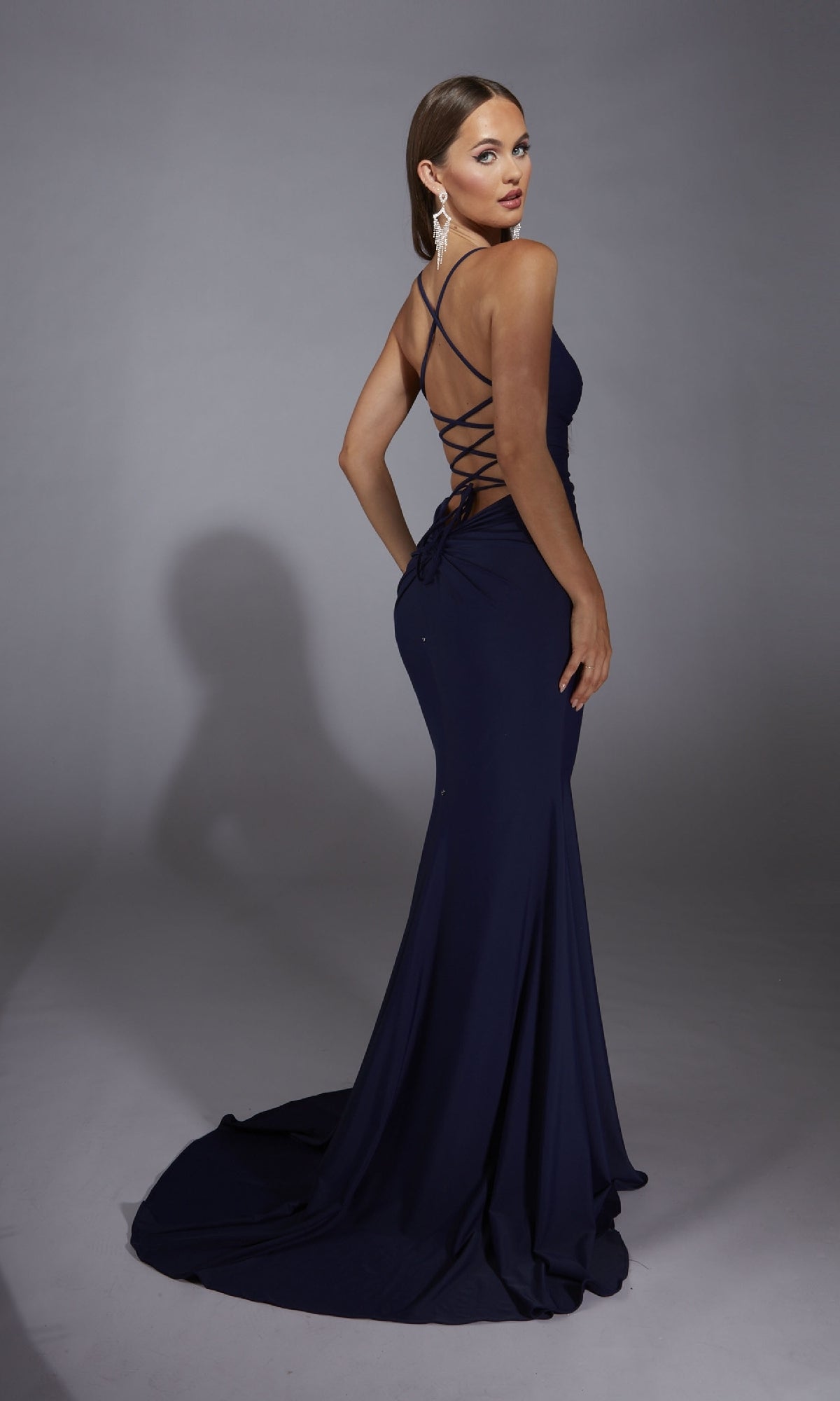 A woman stands on a plain gray background, looking over her shoulder, wearing the Aerie Couture 346 Cowl-Neck Long Prom Dress in navy blue with a floor-length skirt and lace-up strappy back.