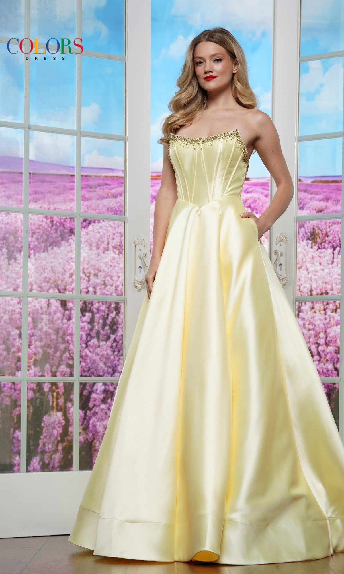 A woman stands indoors by glass doors, wearing the elegant Strapless Corset Prom Ball Gown: Colors Dress 3478 in yellow satin with pockets. Purple flowers bloom outside in the background.