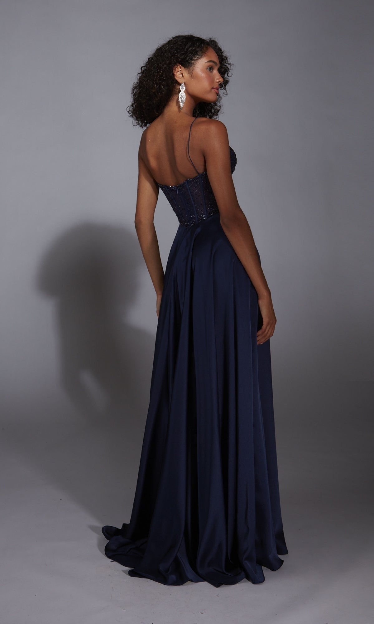 A woman faces away in the Long A-Line Prom Dress: Aerie Couture 348, featuring navy blue fabric, spaghetti straps, a fitted bodice, and a subtle leg slit, set against a plain gray background.