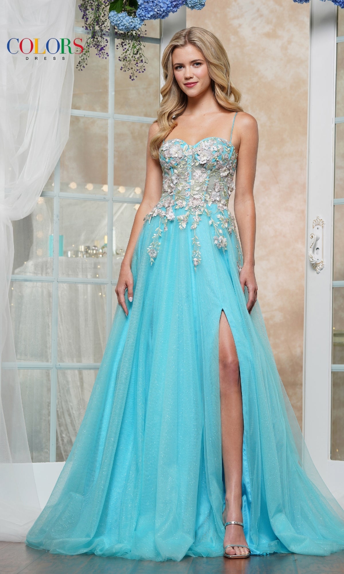 A woman wears the Pastel Long Prom Ball Gown: Colors Dress 3496, a light blue sleeveless evening gown with floral embroidery and a high side slit, standing indoors in front of a glass door.