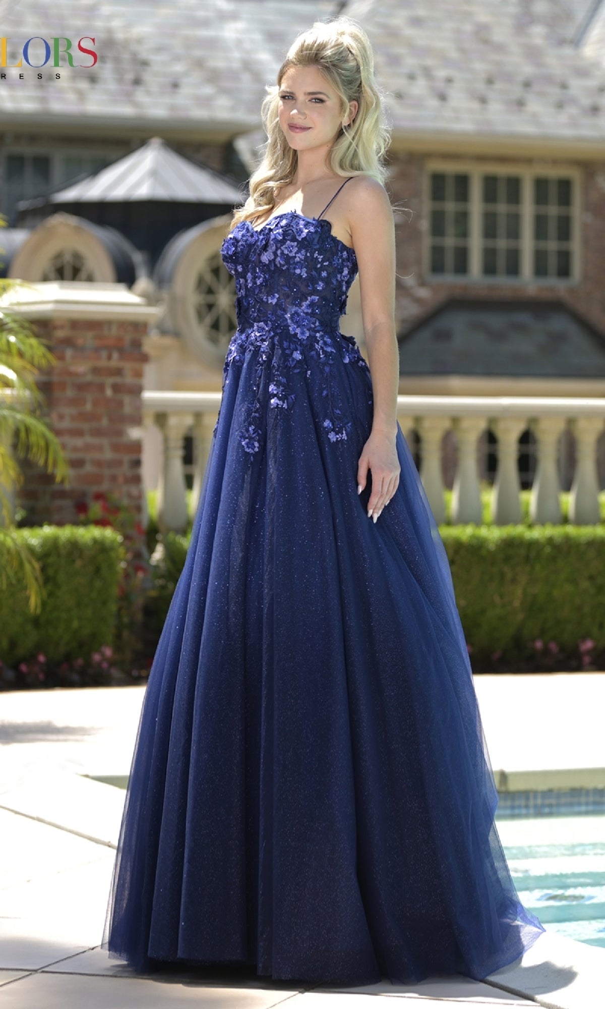 A woman stands outdoors by a pool and large house, wearing the Pastel Long Prom Ball Gown: Colors Dress 3496 with floral detailing on the bodice and thin straps.
