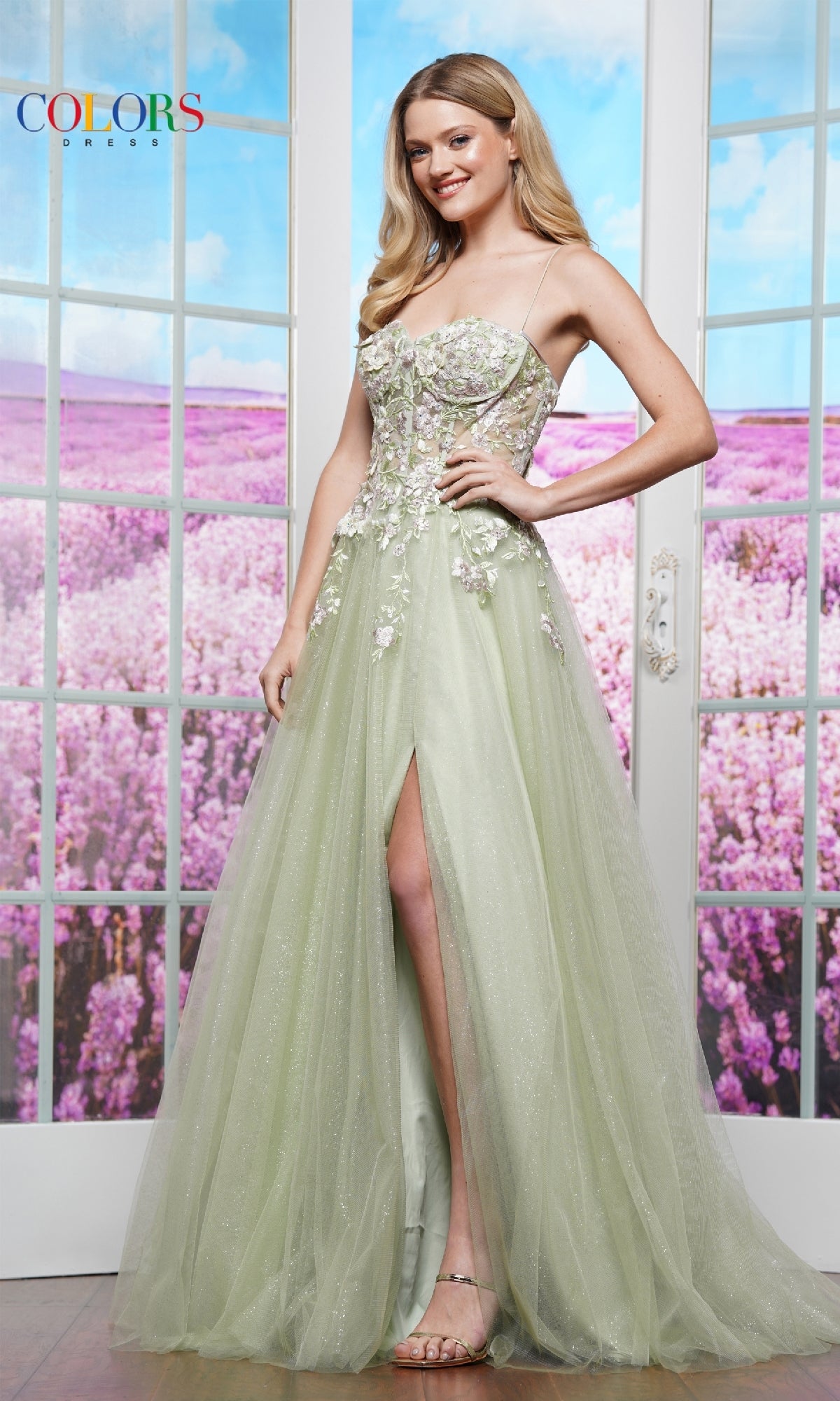A woman in the Pastel Long Prom Ball Gown: Colors Dress 3496—featuring light green fabric, lace-up back, floral adornments, and a thigh-high slit—stands indoors by glass doors, gazing at a lavender field.