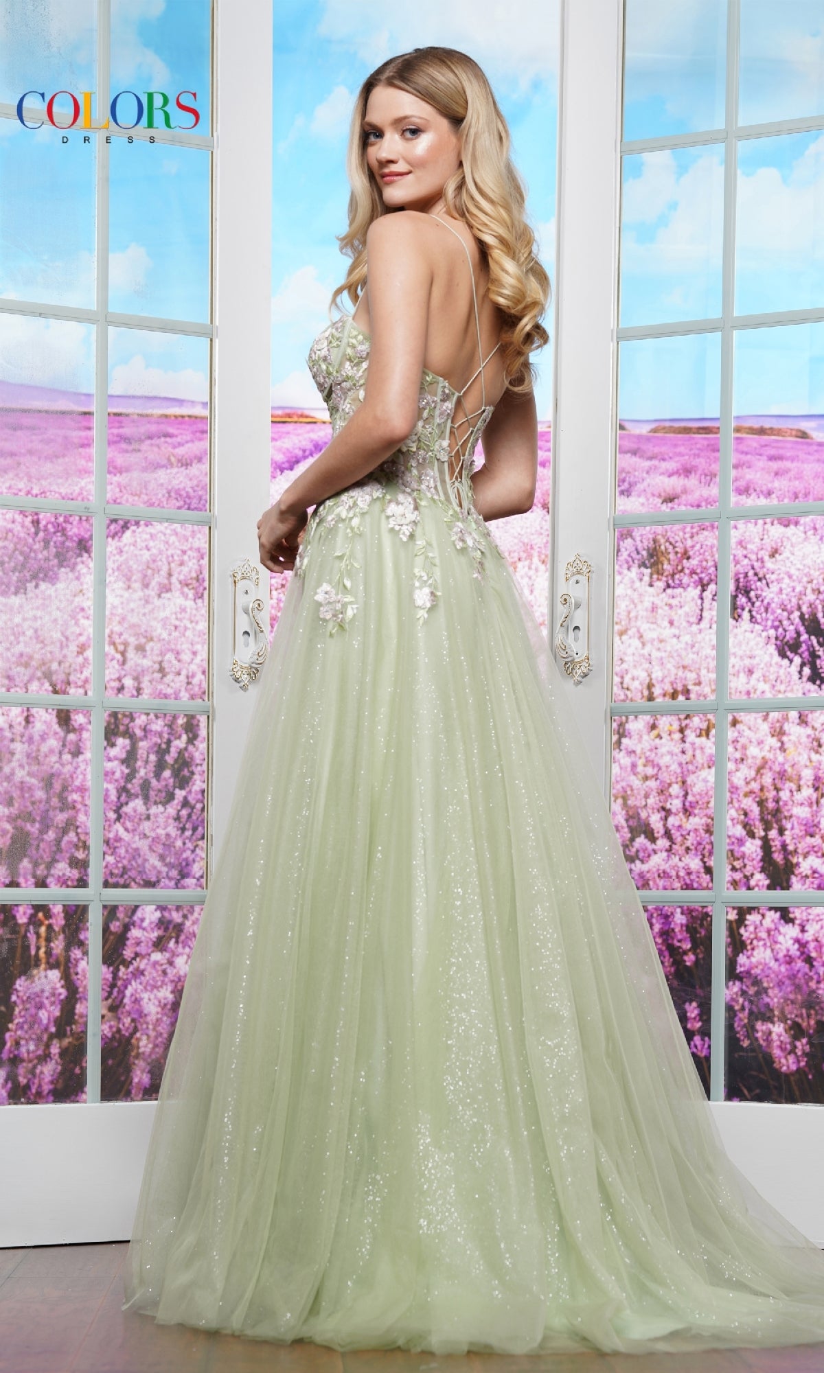 A woman stands in front of glass doors, wearing the Colors Dress 3496—an elegant pastel long prom ball gown—with a field of purple flowers visible outside.