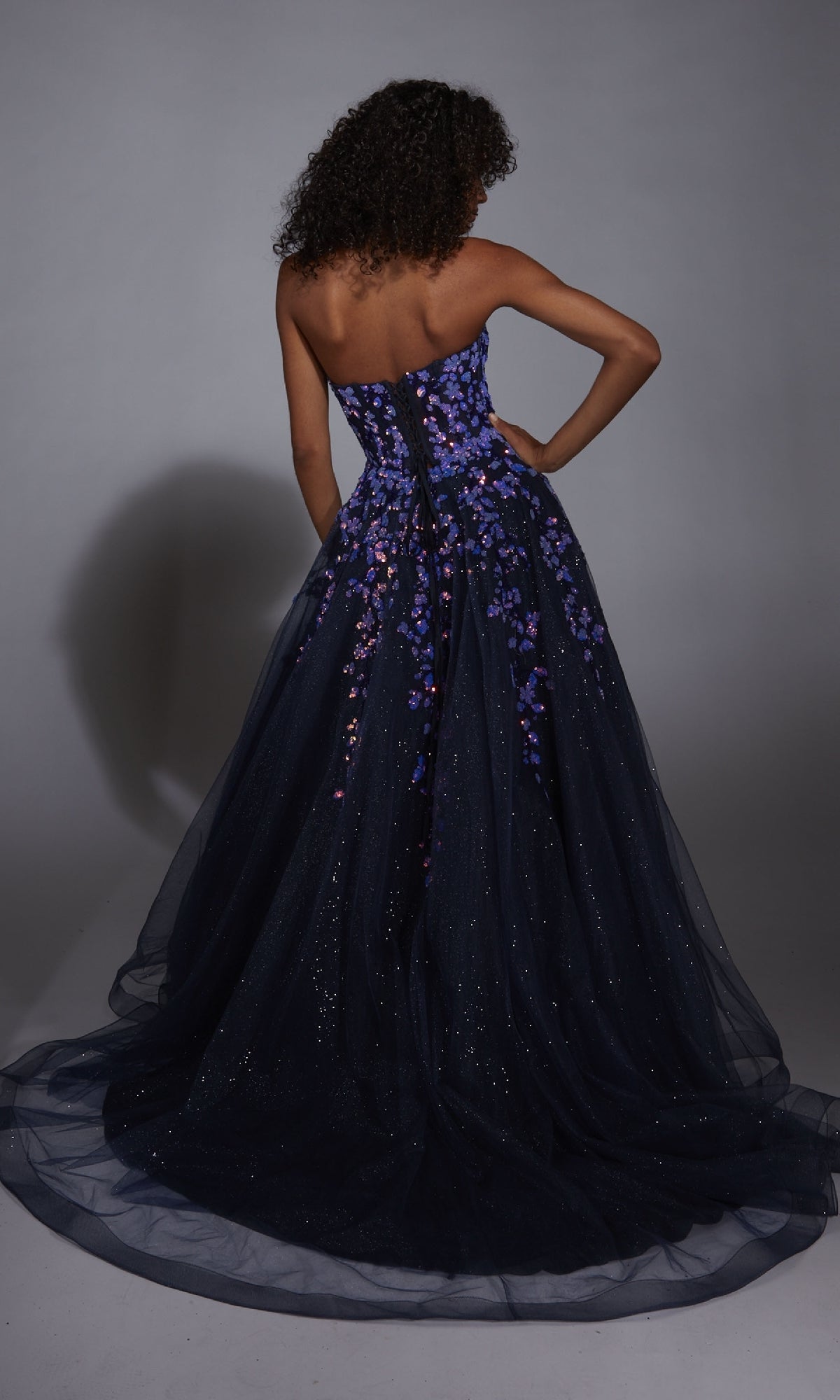 A woman faces away from the camera in the Aerie Couture 350 navy blue prom ball gown, featuring a strapless fitted bodice with purple floral appliques and a voluminous, glittery A-line tulle skirt.