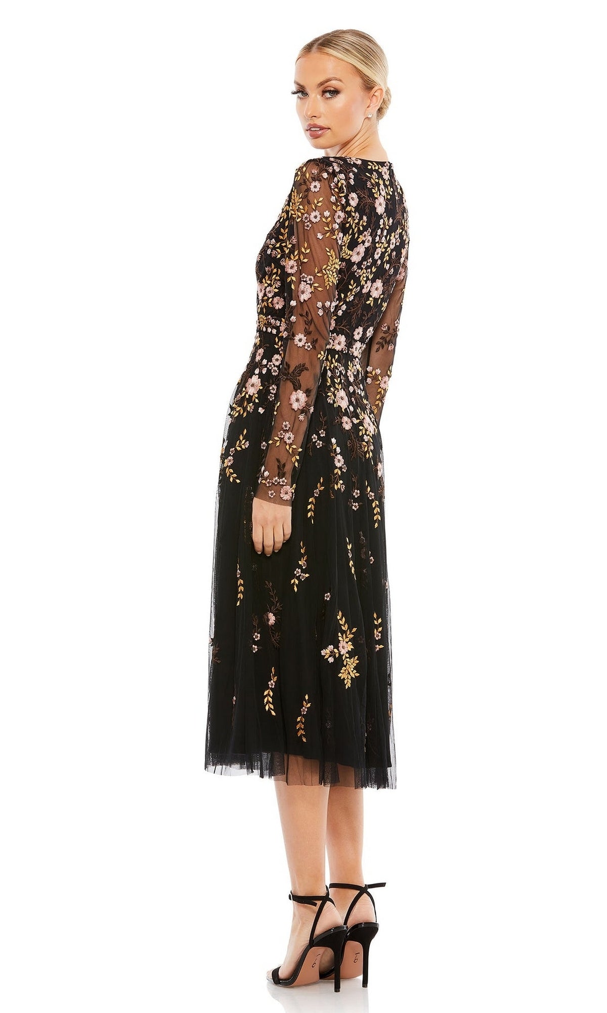 A woman in the Midi Floral Wedding Guest Dress by Mac Duggal, featuring black long sleeves and floral embroidery, stands in high-heeled sandals and looks over her shoulder against a plain white background.