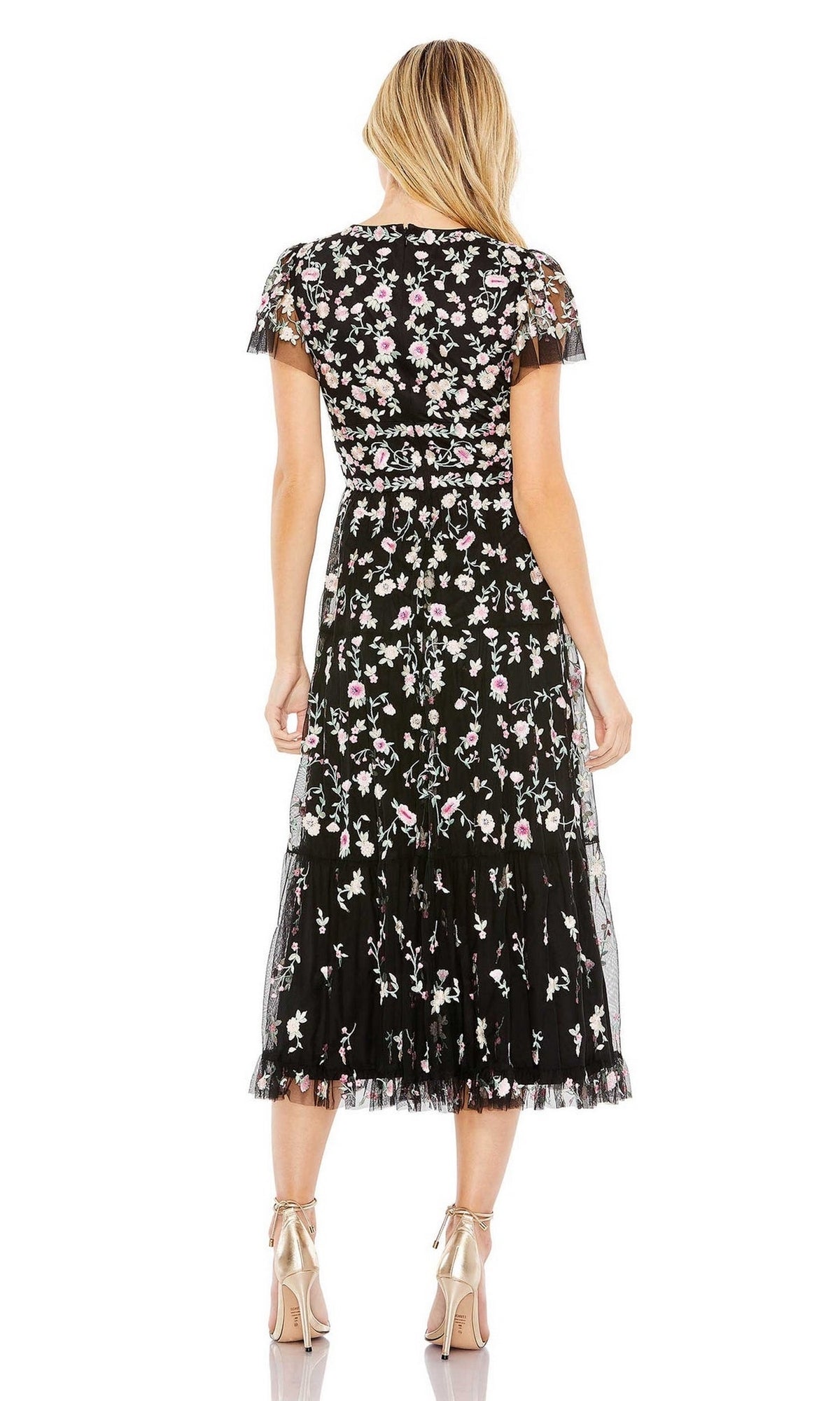 A woman faces away, wearing the Mac Duggal 35104 embroidered wedding-guest dress—a black floral midi with short sleeves—paired with metallic high-heeled sandals.
