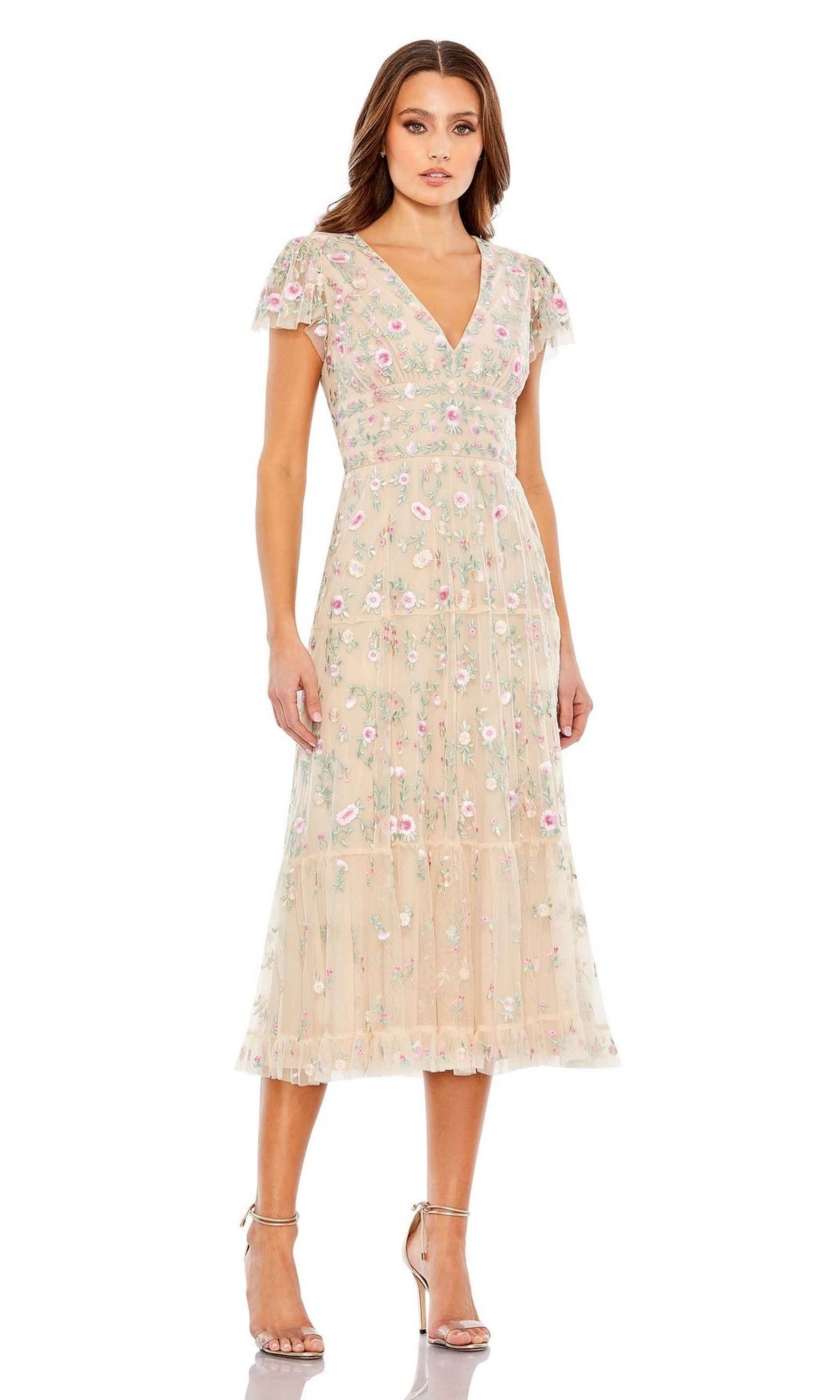 A woman models the Mac Duggal 35104 Embroidered Wedding-Guest Dress in light beige with floral embroidery, short sleeves, and a V-neckline. She pairs it with neutral heels against a white background.