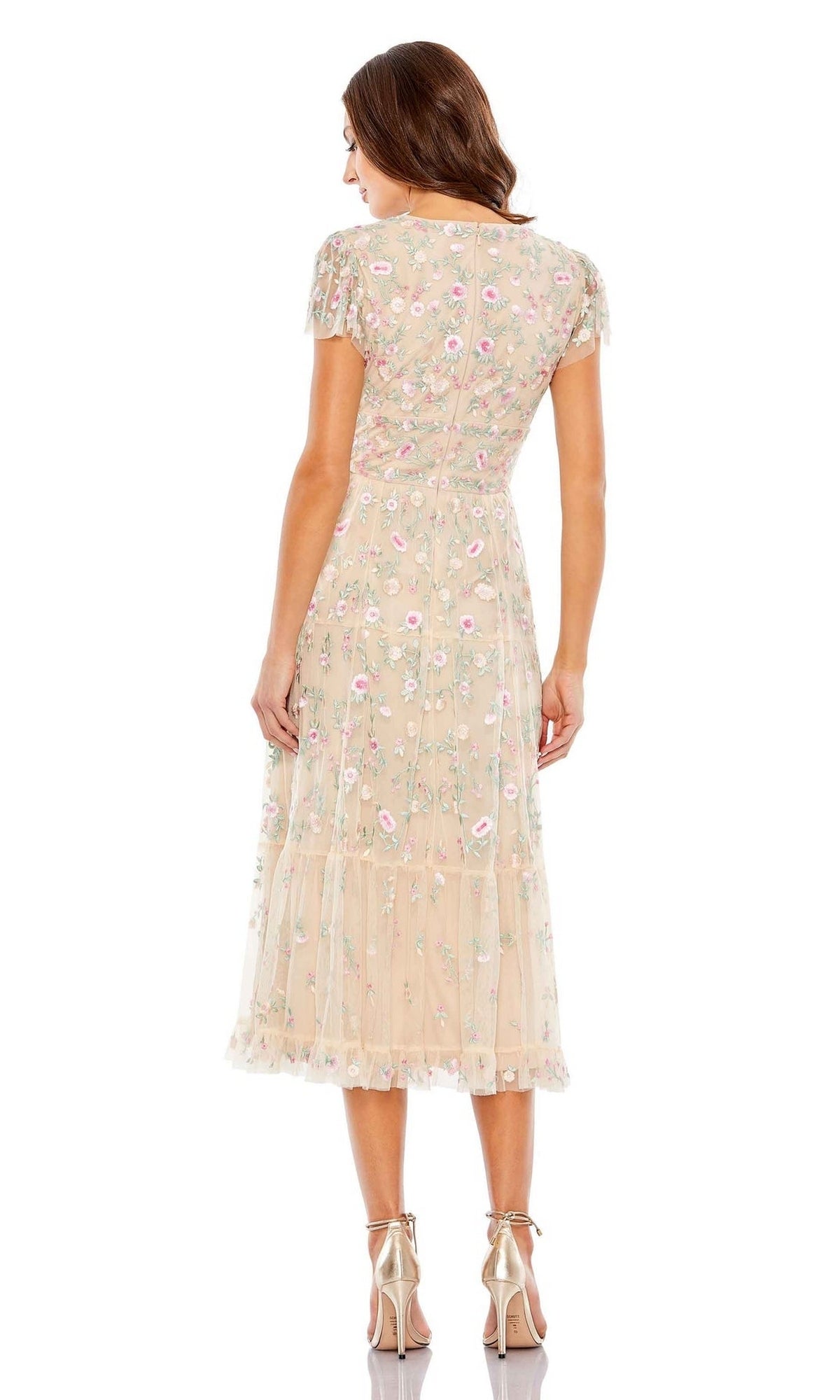 A woman faces away, wearing the Mac Duggal 35104 beige, ankle-length embroidered wedding-guest dress with short sleeves, paired with metallic high-heeled sandals.