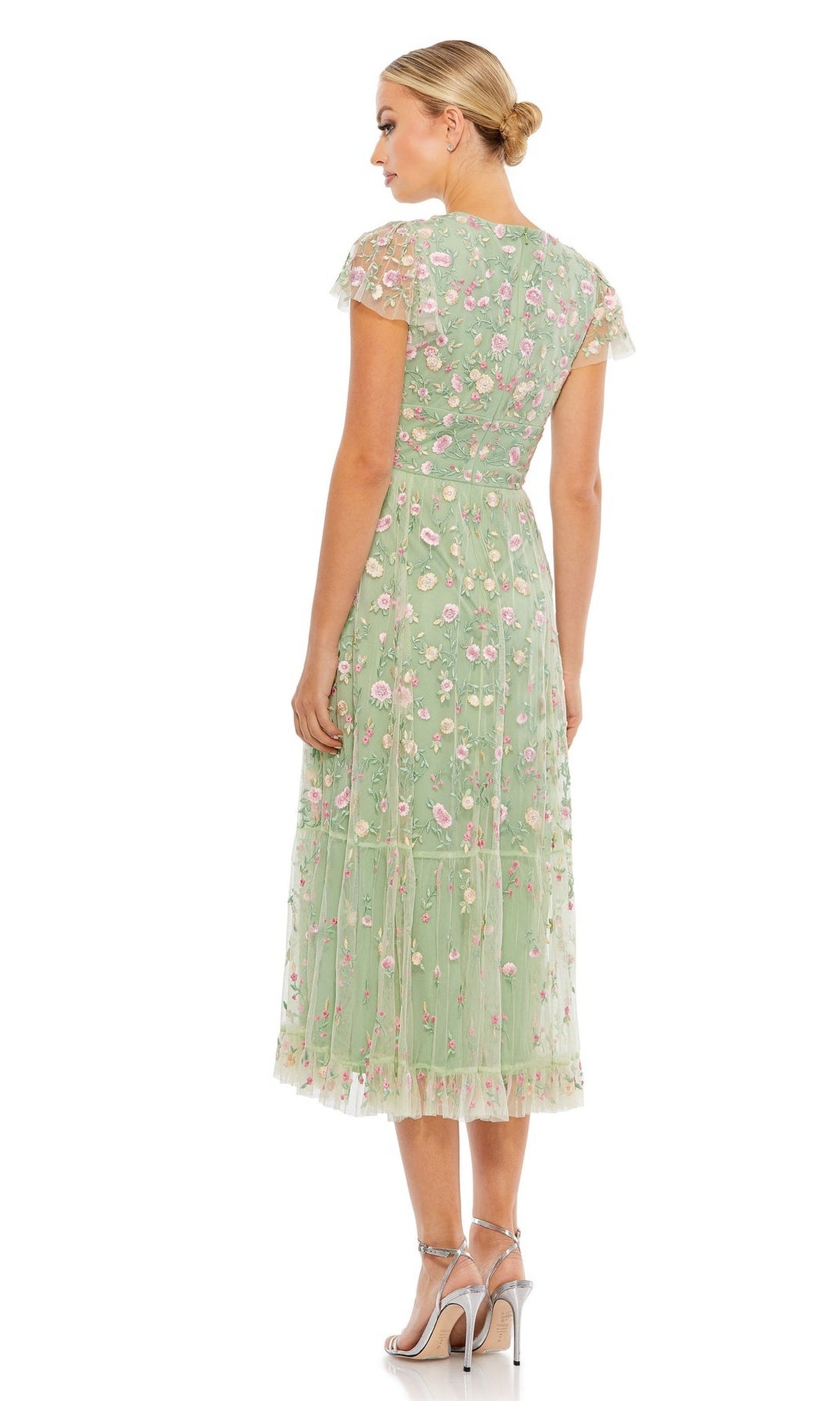 A woman stands facing away, wearing the Mac Duggal 35104 Embroidered Wedding-Guest Dress in light green with pink floral accents and silver high-heeled sandals, set against a white background.