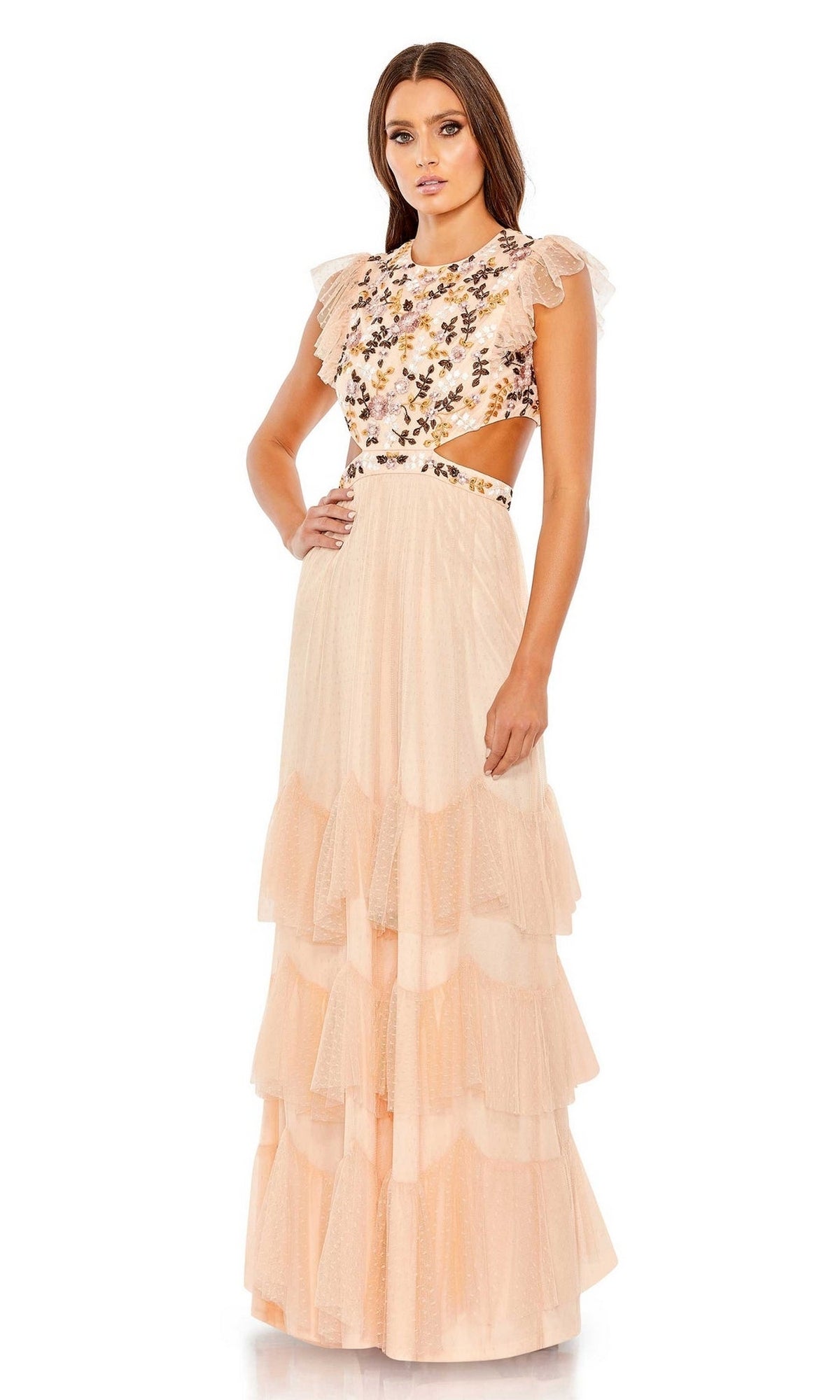 A woman models the Long Formal Dress 35106 by Mac Duggal—a floor-length, tiered gown featuring floral embroidery on the bodice, cap sleeves, and cutout sides. She stands facing forward with one hand on her hip.