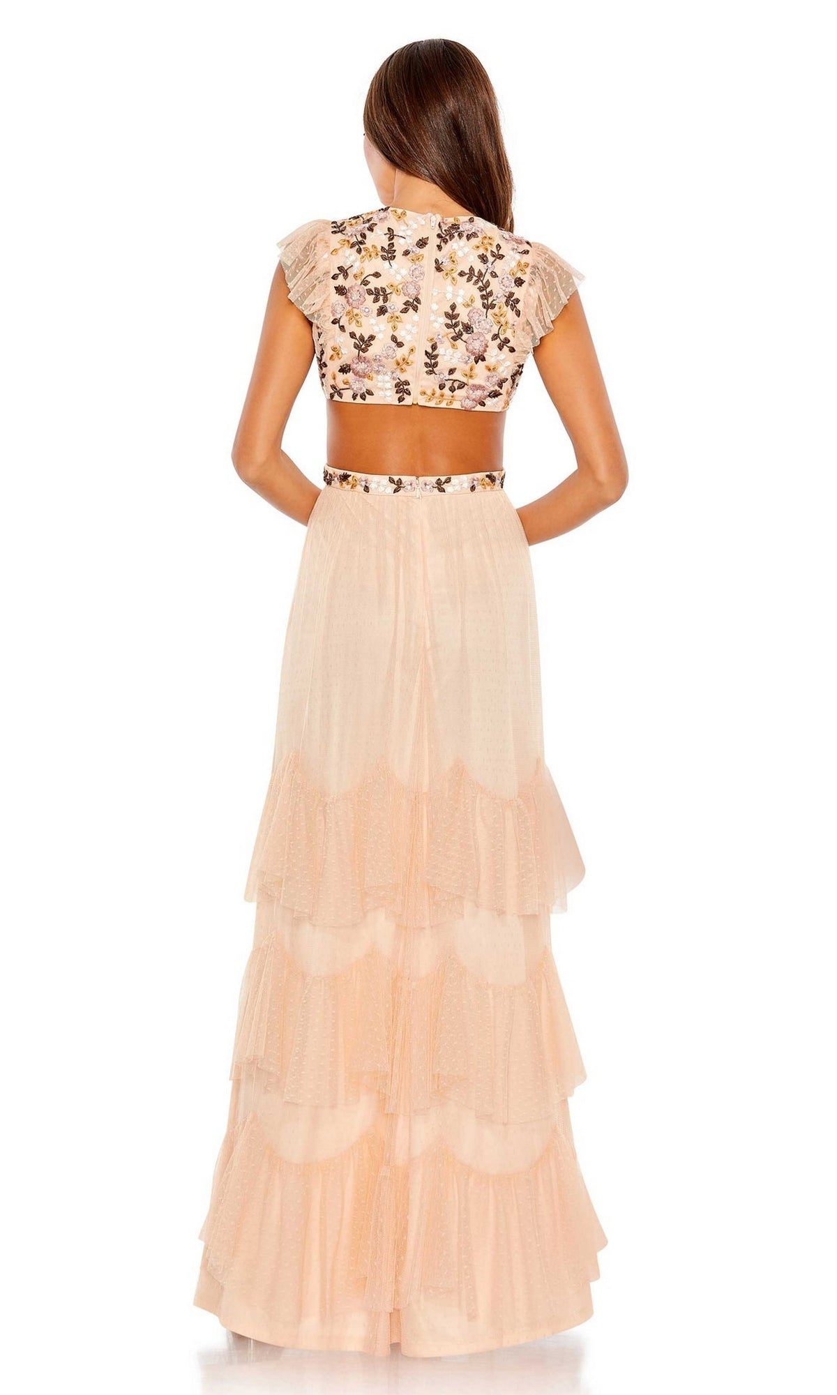 A woman stands facing away, wearing the Long Formal Dress 35106 by Mac Duggal—a peach-toned tiered gown with a short-sleeved, floral embroidered crop top featuring delicate designs.