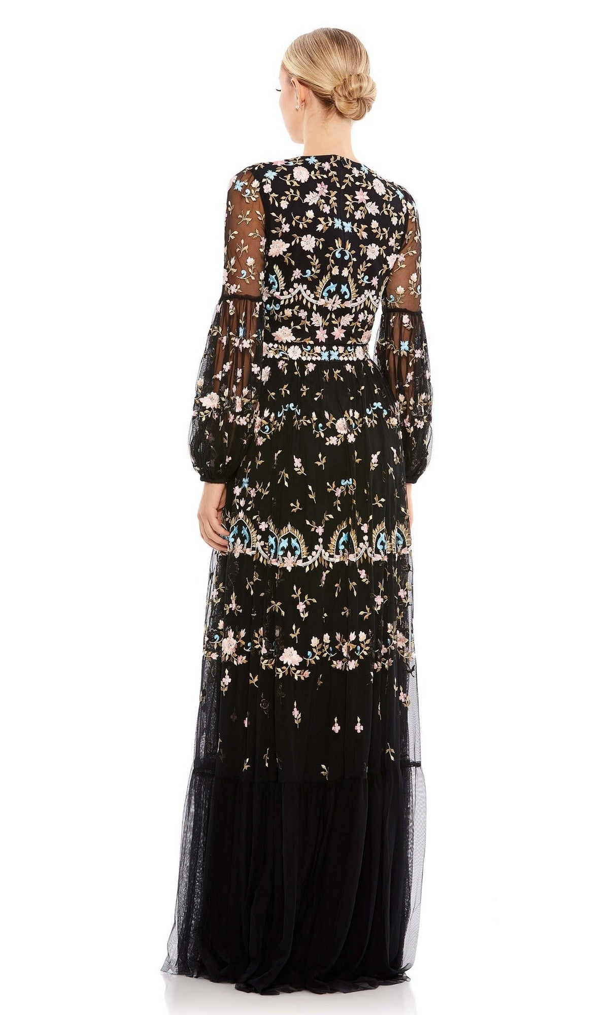 A woman stands facing away, wearing the Mac Duggal Long Formal Dress 35111 with sheer long sleeves and floral embroidery, her hair styled in a neat low bun.