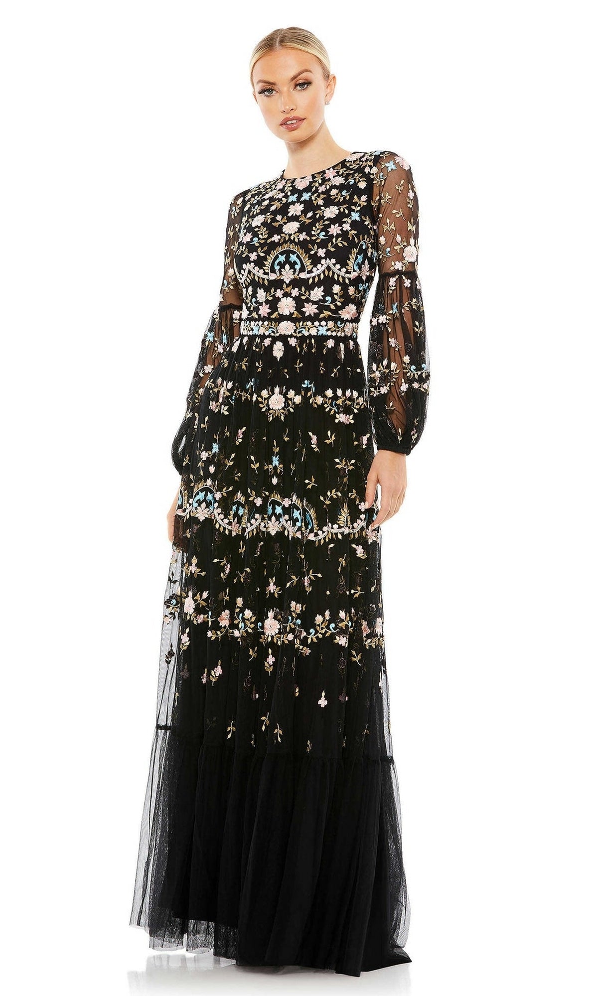 A woman wears the Long Formal Dress 35111 by Mac Duggal—a black floor-length gown with colorful floral embroidery, sheer long sleeves, and a fitted bodice.