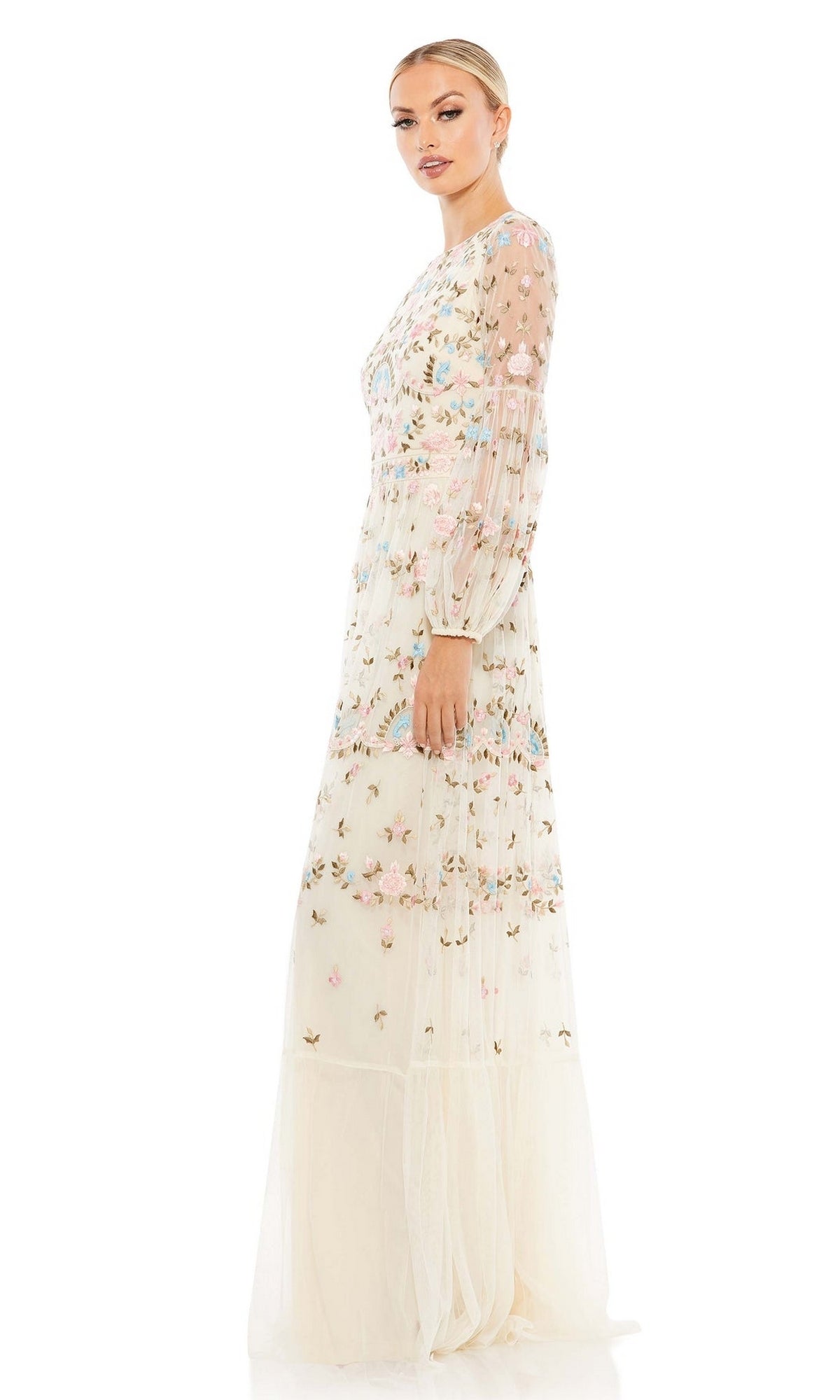A woman stands sideways against a white background wearing the Long Formal Dress 35111 by Mac Duggal, a sheer, cream-colored gown with pastel floral embroidery.