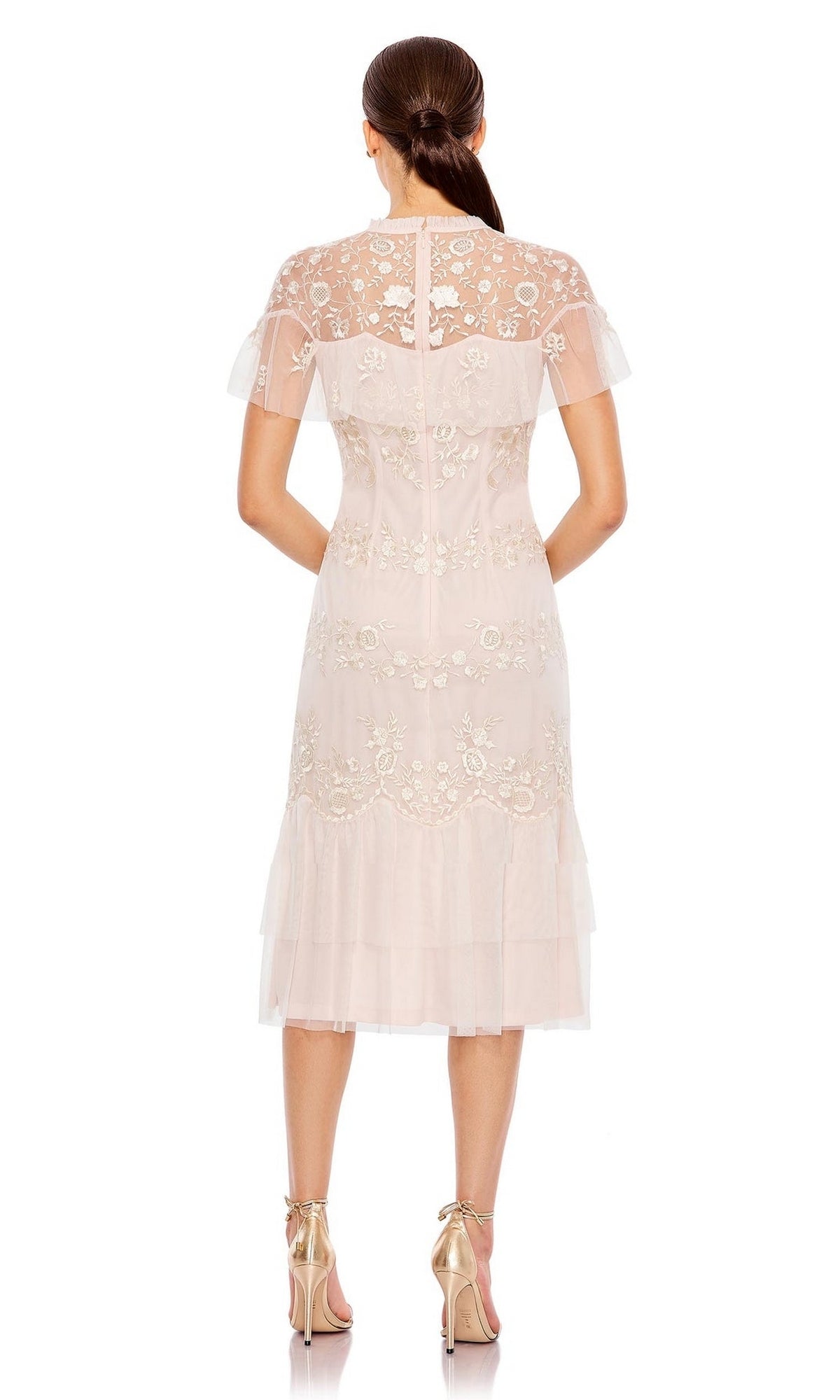 A woman faces away, wearing the Short Wedding Guest Dress 35147 by Mac Duggal—a pale pink, knee-length lace dress with floral embroidery and a layered hem—paired with gold high-heeled sandals.
