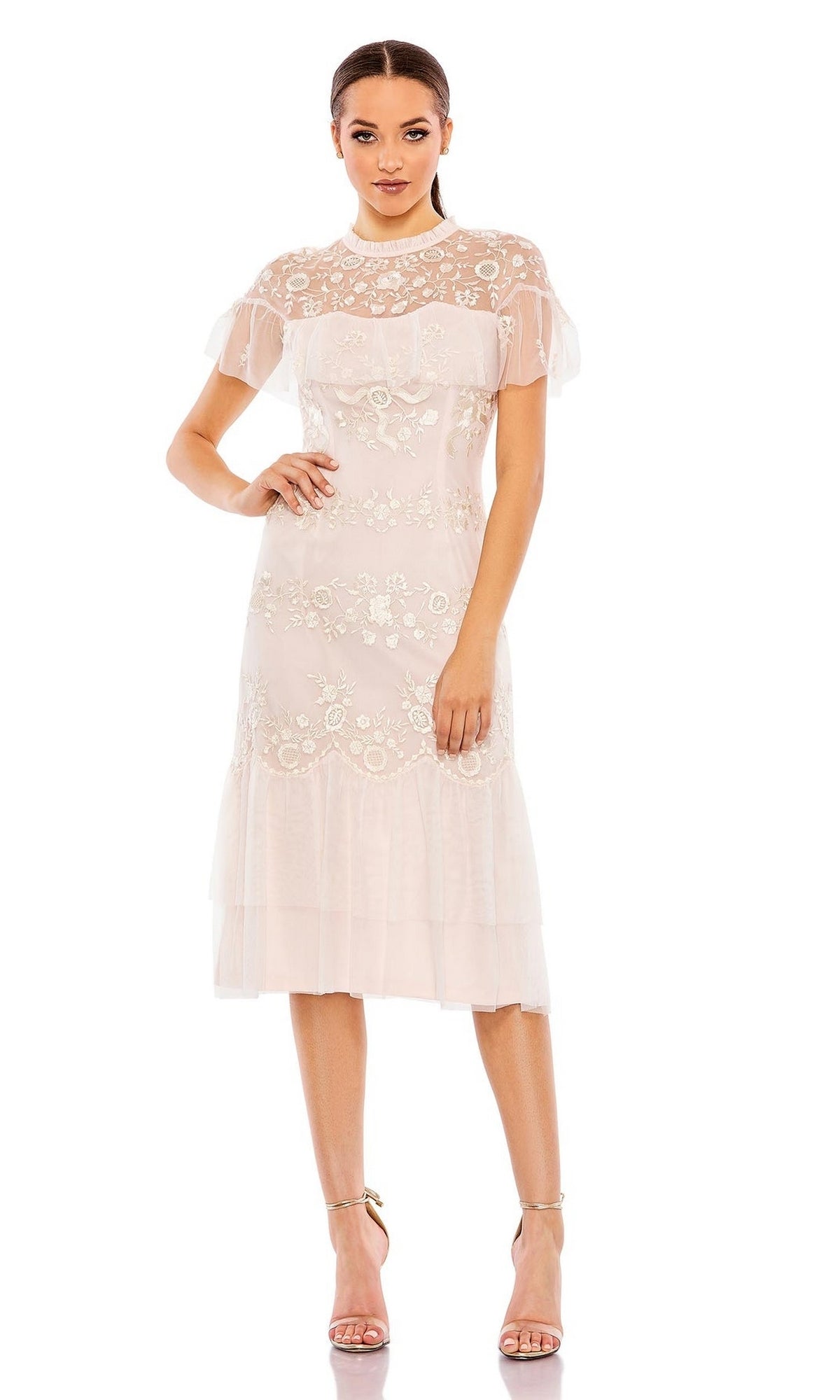 A woman stands in heels against a white background wearing the Mac Duggal Short Wedding Guest Dress 35147—a light pink, knee-length dress with floral lace details, short sleeves, and a sheer overlay that blends classic style with modern charm.
