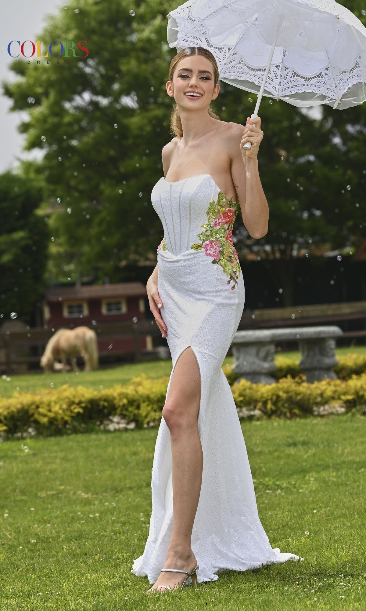 A woman in a Sequin Strapless Prom Dress: Colors Dress 3536 holds a lace parasol while standing on grass, with benches, bushes, and a horse in the background.