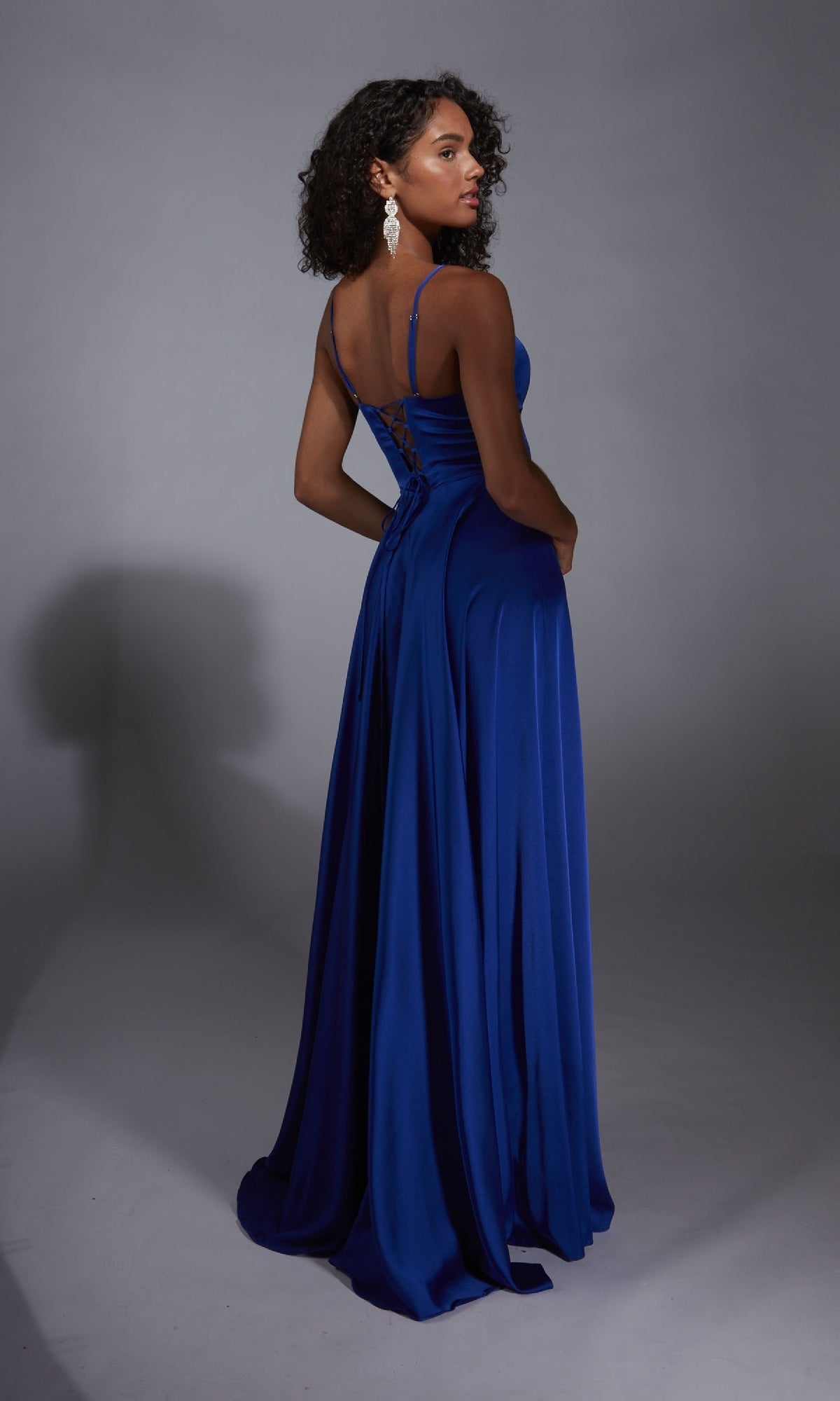 A woman faces away, modeling the Aerie Couture 354 long prom dress in royal blue satin, featuring a lace-up back and floor-length A-line silhouette, against a plain gray background.