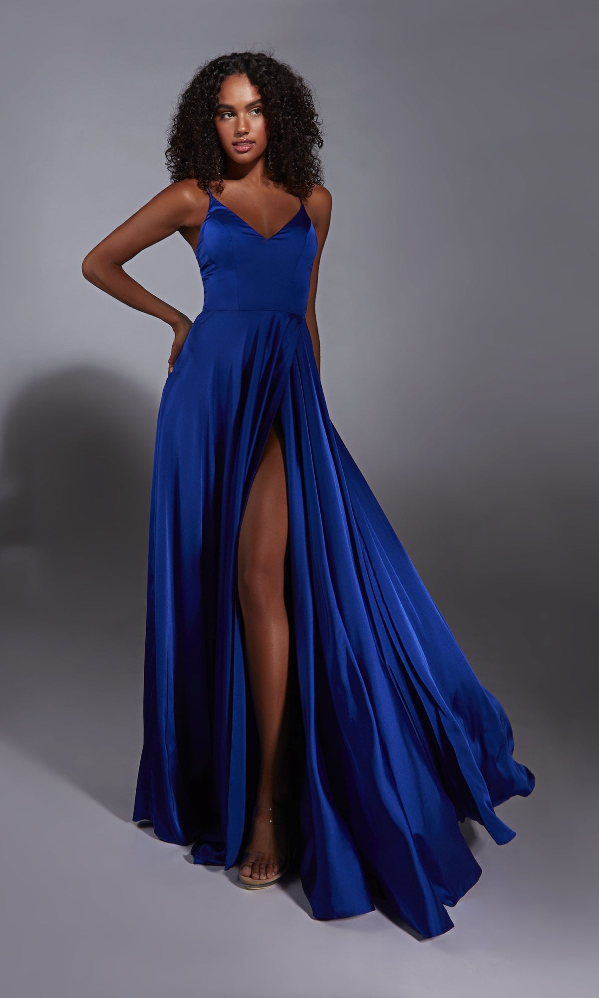 A woman poses against a gray background wearing the Aerie Couture 354 long prom dress in royal blue, featuring a sleeveless satin design with an A-line silhouette and deep leg slit, her hand resting on her hip.