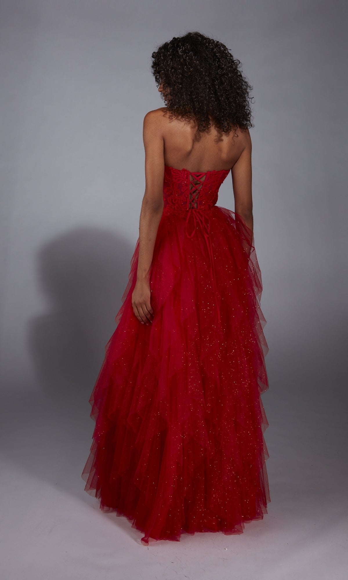 A woman with curly hair models the A-Line Glitter Prom Dress: Aerie Couture 356, which features a strapless design, lace corset bodice, and ruffled skirt, standing against a plain background.