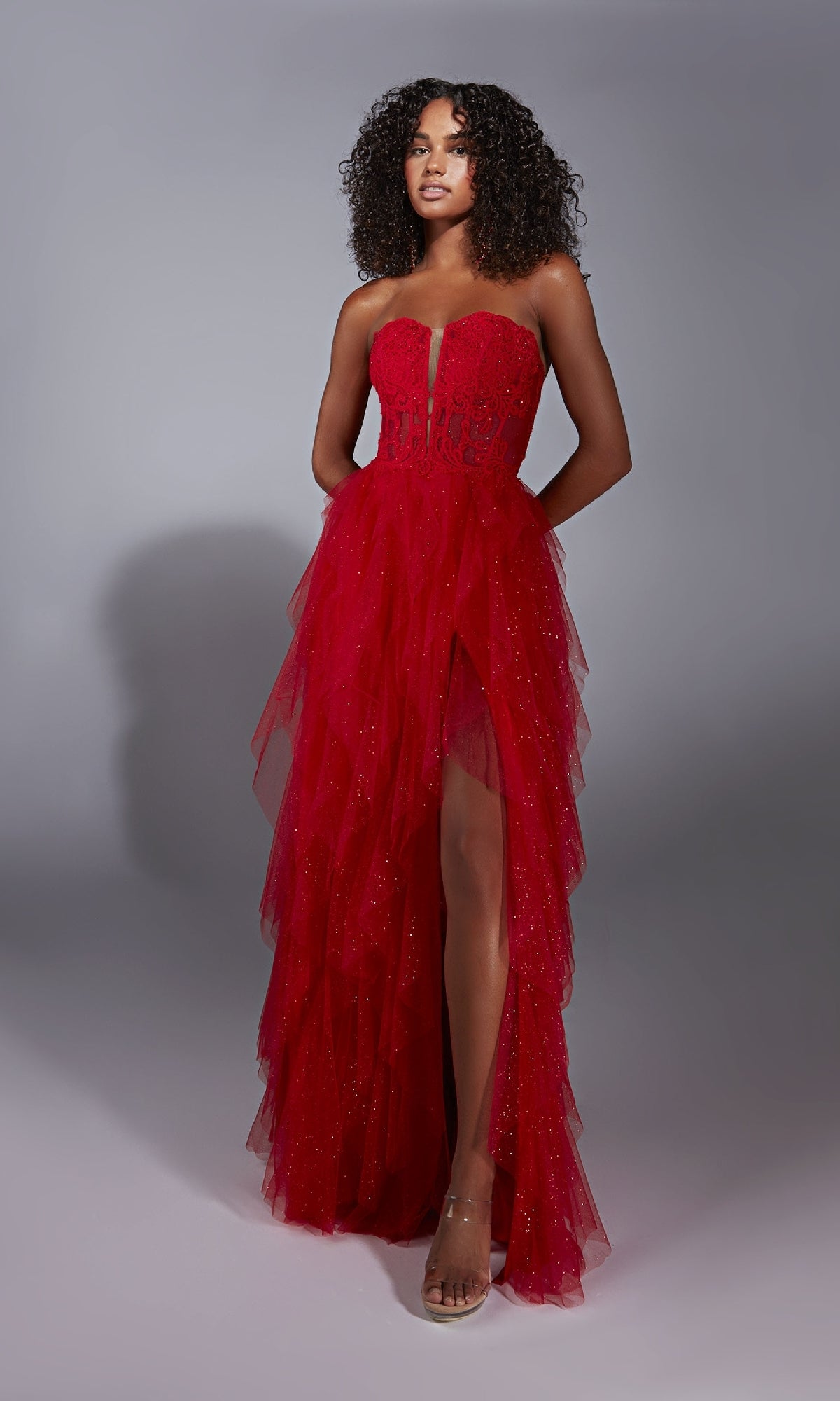 A woman poses in the A-Line Glitter Prom Dress: Aerie Couture 356, showcasing a strapless design with a sheer bodice, ruffled A-line skirt, sparkling details, and a high front slit against a plain gray background.