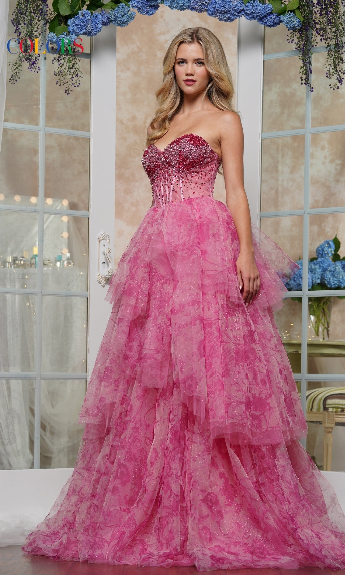 A woman stands indoors in front of mirrored doors with blue flowers, wearing the Strapless Print Prom Ball Gown: Colors Dress 3572, featuring a beaded corset bodice and a layered pink skirt.
