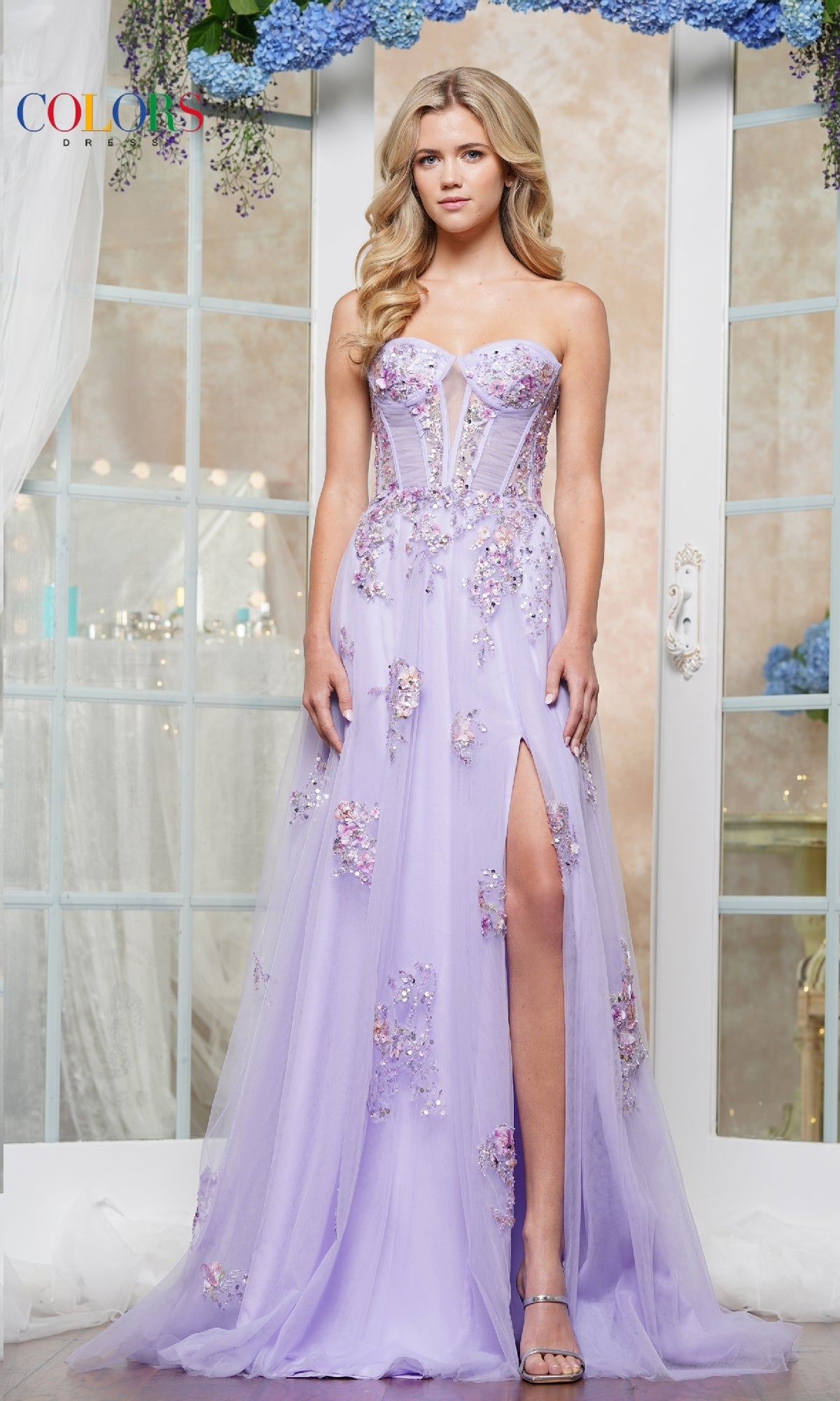 A woman poses indoors in front of floral-adorned French doors, wearing the Strapless Long Prom Ball Gown: Colors Dress 3575—a lilac gown with floral appliqué details and a thigh-high slit.