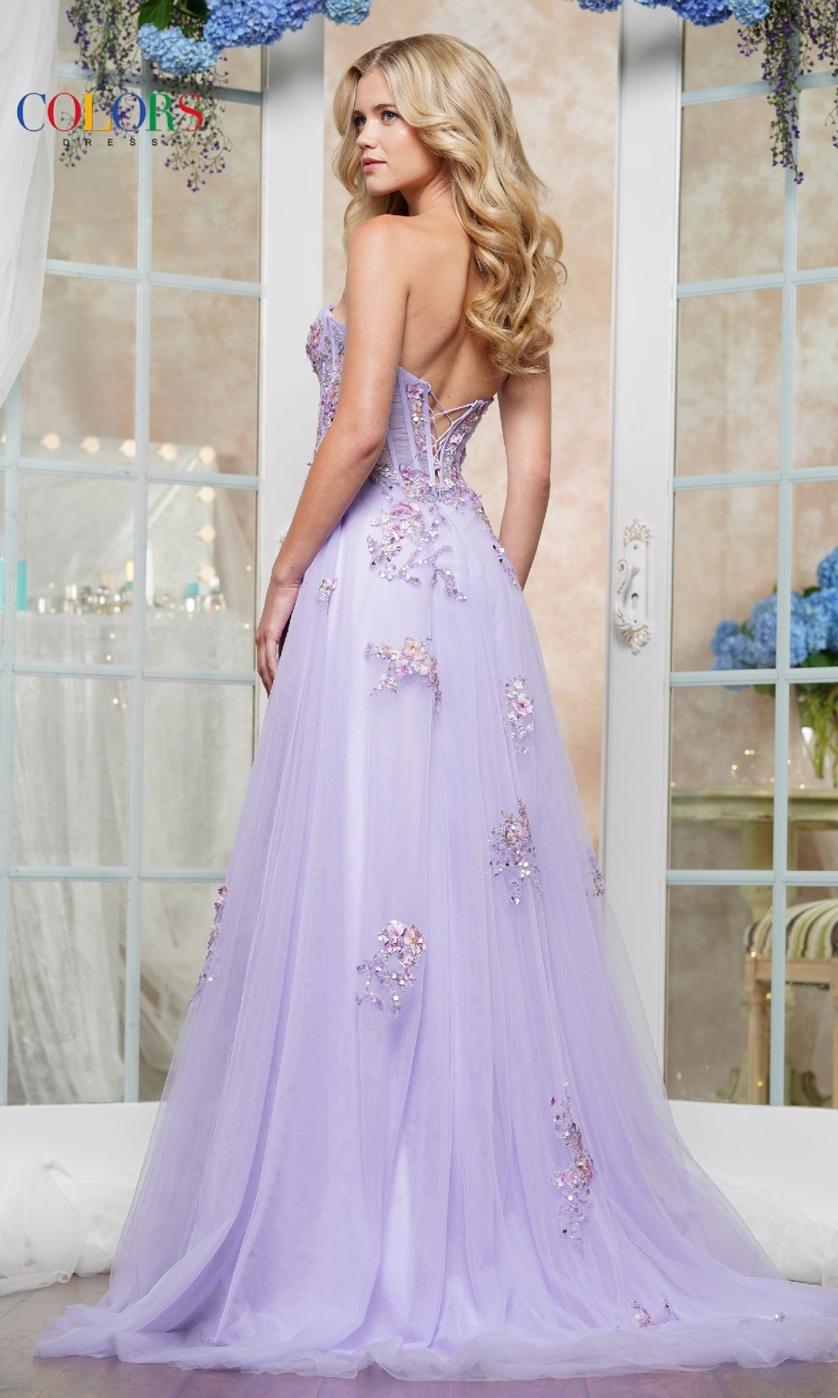 A woman poses indoors in the Strapless Long Prom Ball Gown: Colors Dress 3575, a lavender gown with floral embellishments and an elegant lace-up back.