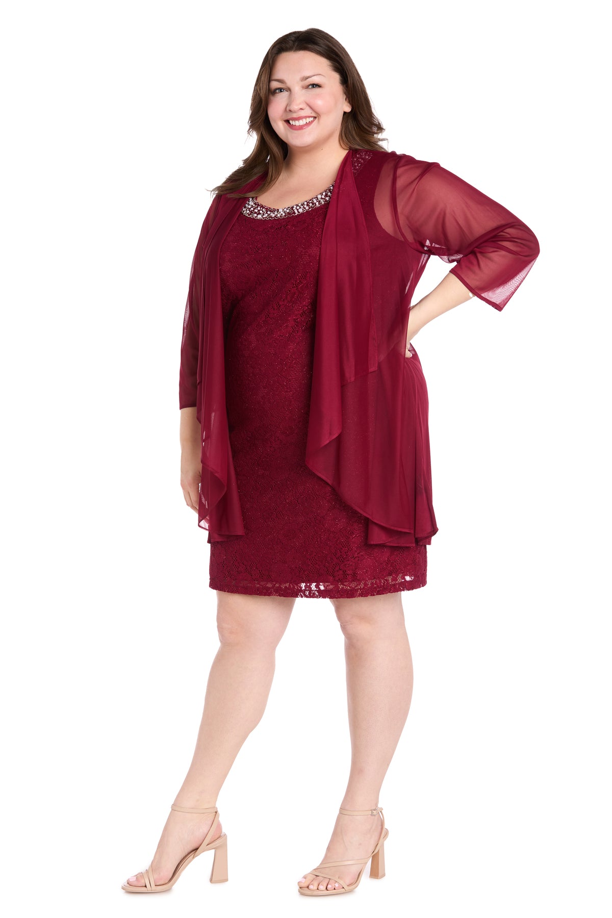 A woman stands smiling against a white background wearing the R&M Richards Plus-Size Short Jacket Dress 3594W, a knee-length lace party dress in burgundy with a matching sheer overlay and nude heels.