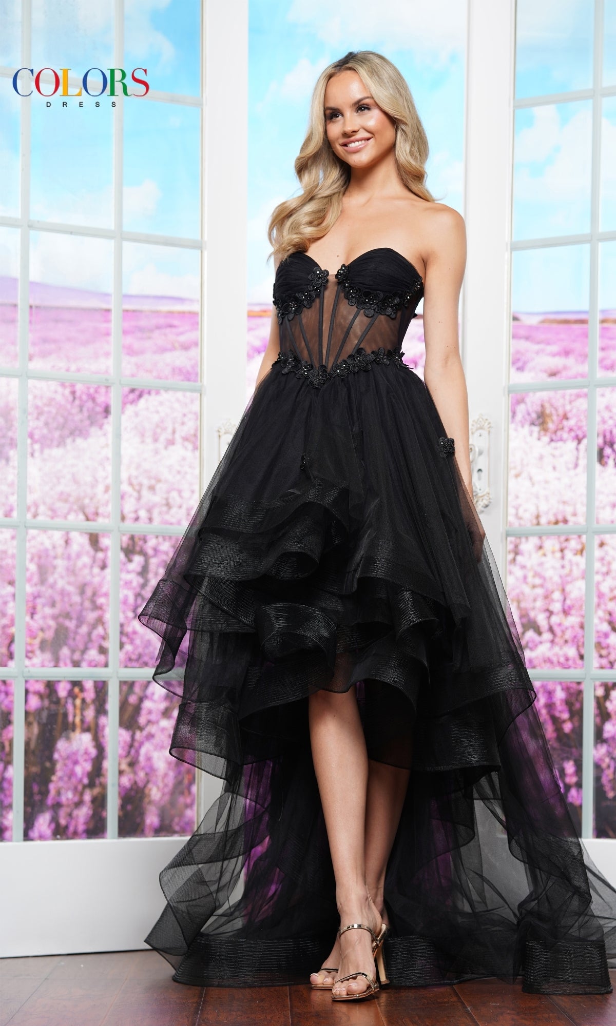 A woman wears the Strapless High-Low Prom Dress: Colors Dress 3600 indoors before glass doors, with purple flowers outside. The elegant black tulle gown offers a charming silhouette perfect for special occasions.