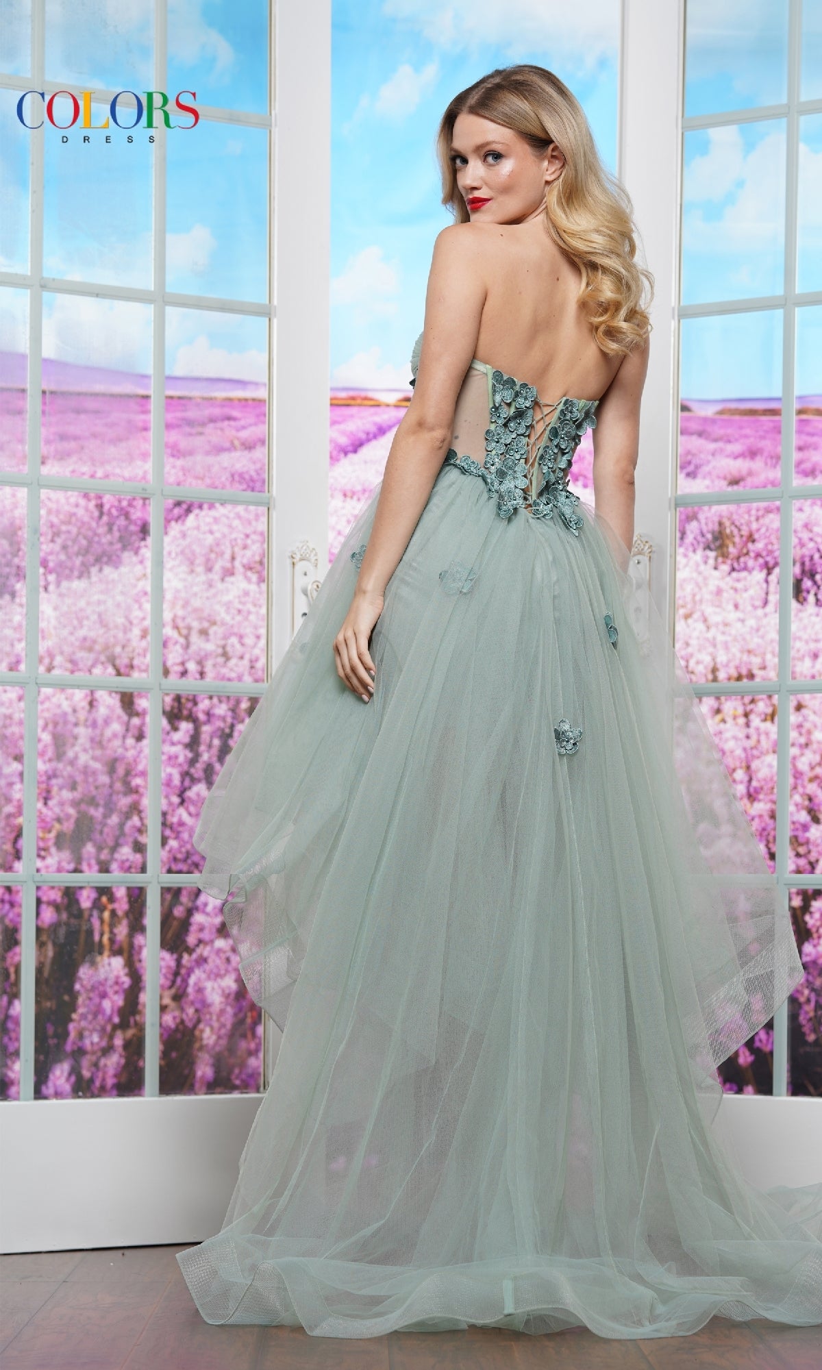 A woman stands in front of glass doors with a field of purple flowers, wearing the Strapless High-Low Prom Dress: Colors Dress 3600 in light green, featuring floral details.