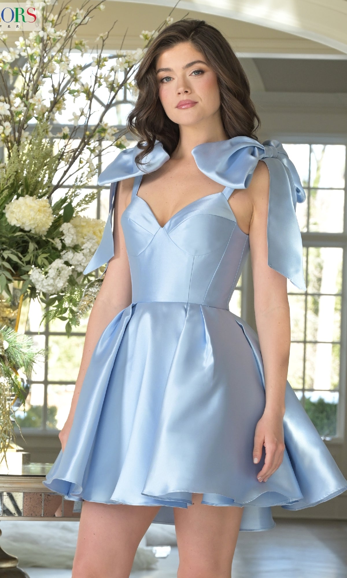 A woman stands indoors near a window and floral arrangement wearing the Shoulder Bow Short Hoco Dress: Colors Dress 3640, a light blue satin babydoll-style dress with large shoulder bows.