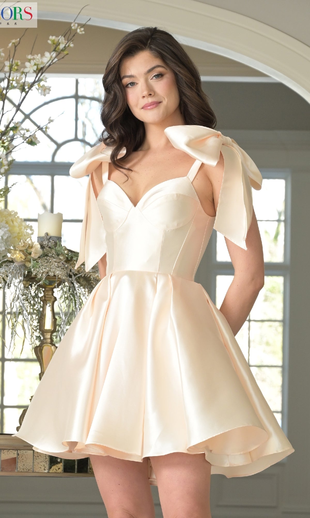 A woman smiles at the camera, standing indoors by a window with her hands in the pockets of the pale pink Shoulder Bow Short Hoco Dress: Colors Dress 3640, featuring a satin sweetheart neckline and large bow straps.