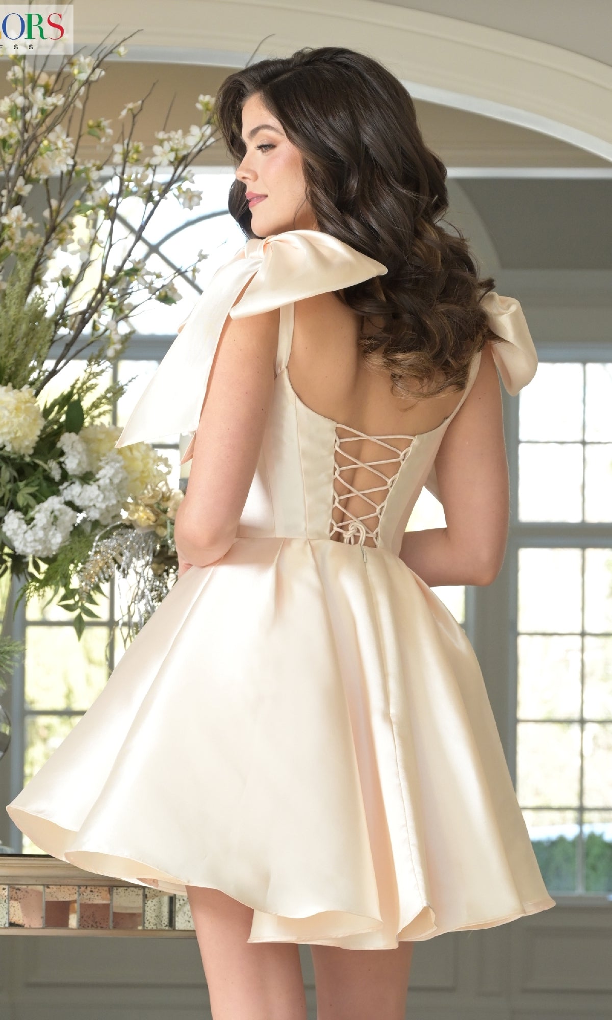 A woman stands indoors by a window and flowers, wearing the Shoulder Bow Short Hoco Dress: Colors Dress 3640. Its light satin fabric, a-line babydoll silhouette, lace-up back, and large shoulder bows create a sweet and stylish look.