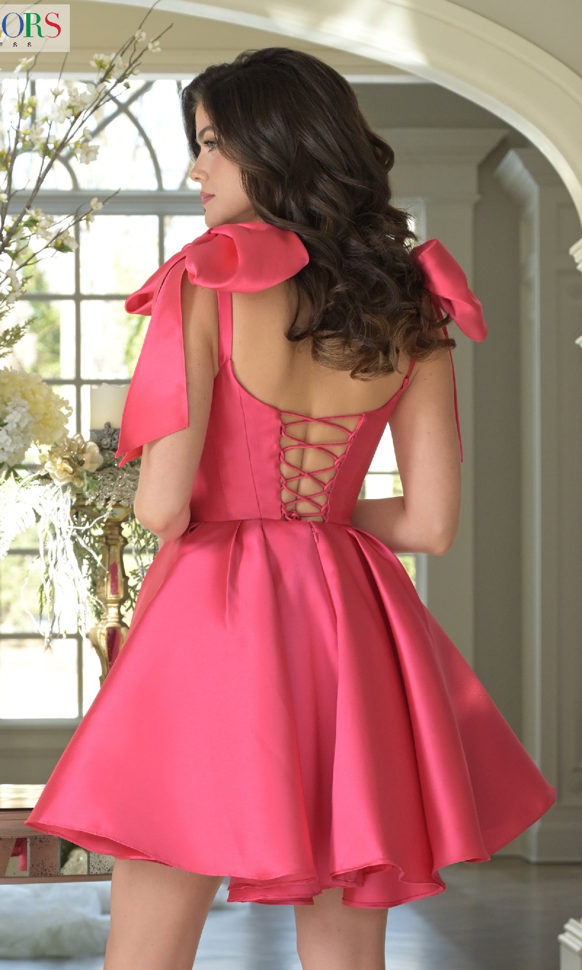A woman stands indoors, facing away, wearing the Shoulder Bow Short Hoco Dress: Colors Dress 3640—a bright pink satin babydoll with ribbon shoulder bows and a lace-up back.