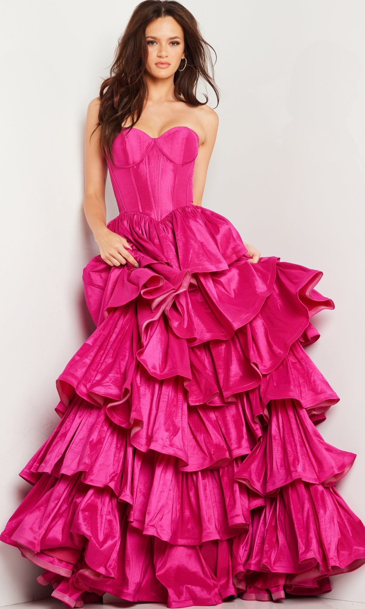 A woman models the Jovani 36619 Strapless Ruffled Prom Ball Gown in bright pink, with a fitted bodice and layered ruffled skirt, standing against a light background.
