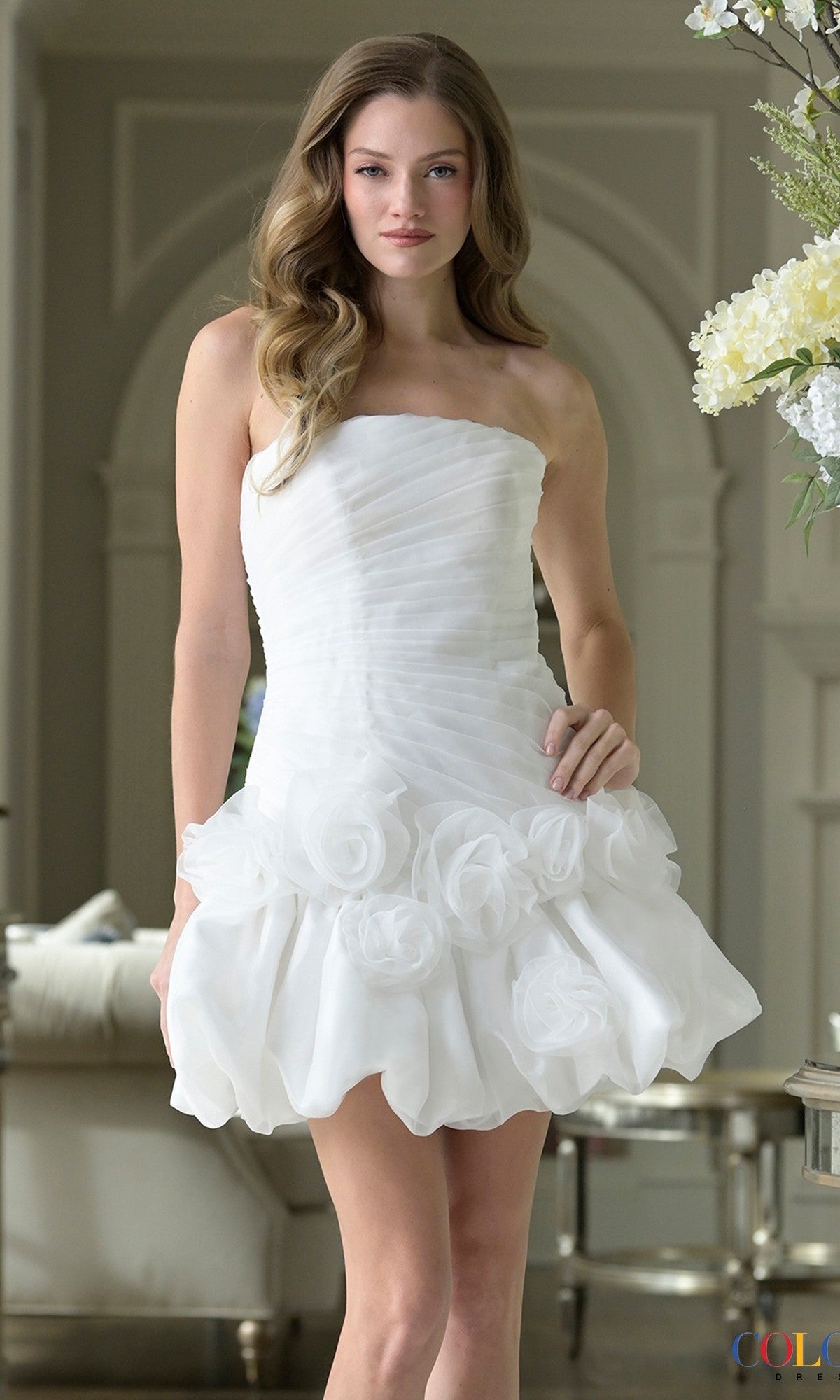 A woman stands indoors in a bright, elegant room wearing the white Strapless Bubble-Hem Hoco Dress: Colors Dress 3671.