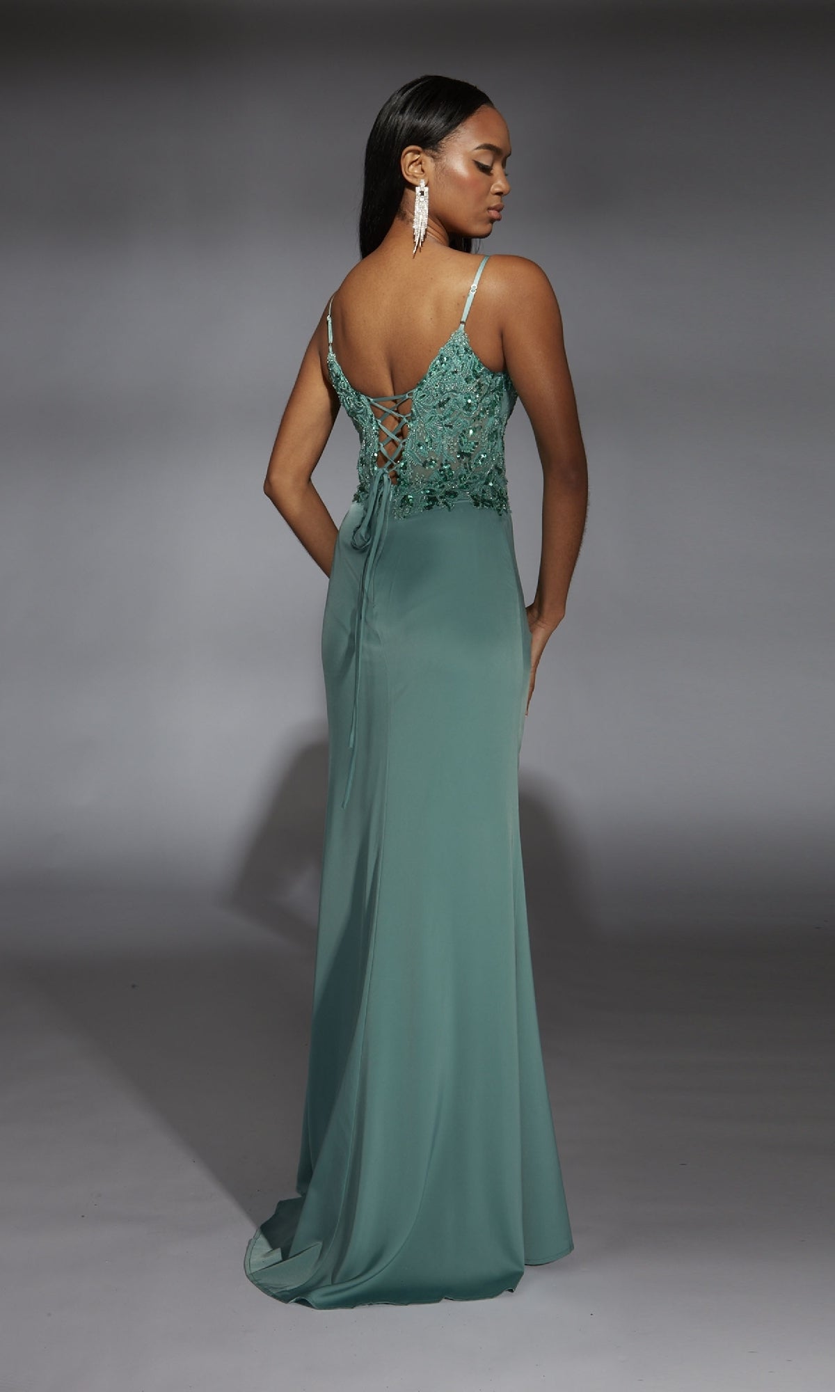 A woman faces away against a gray background, wearing the Aerie Couture 369— a long sage green prom dress with thin straps, a lace-up back, and floral embroidery on the bodice.