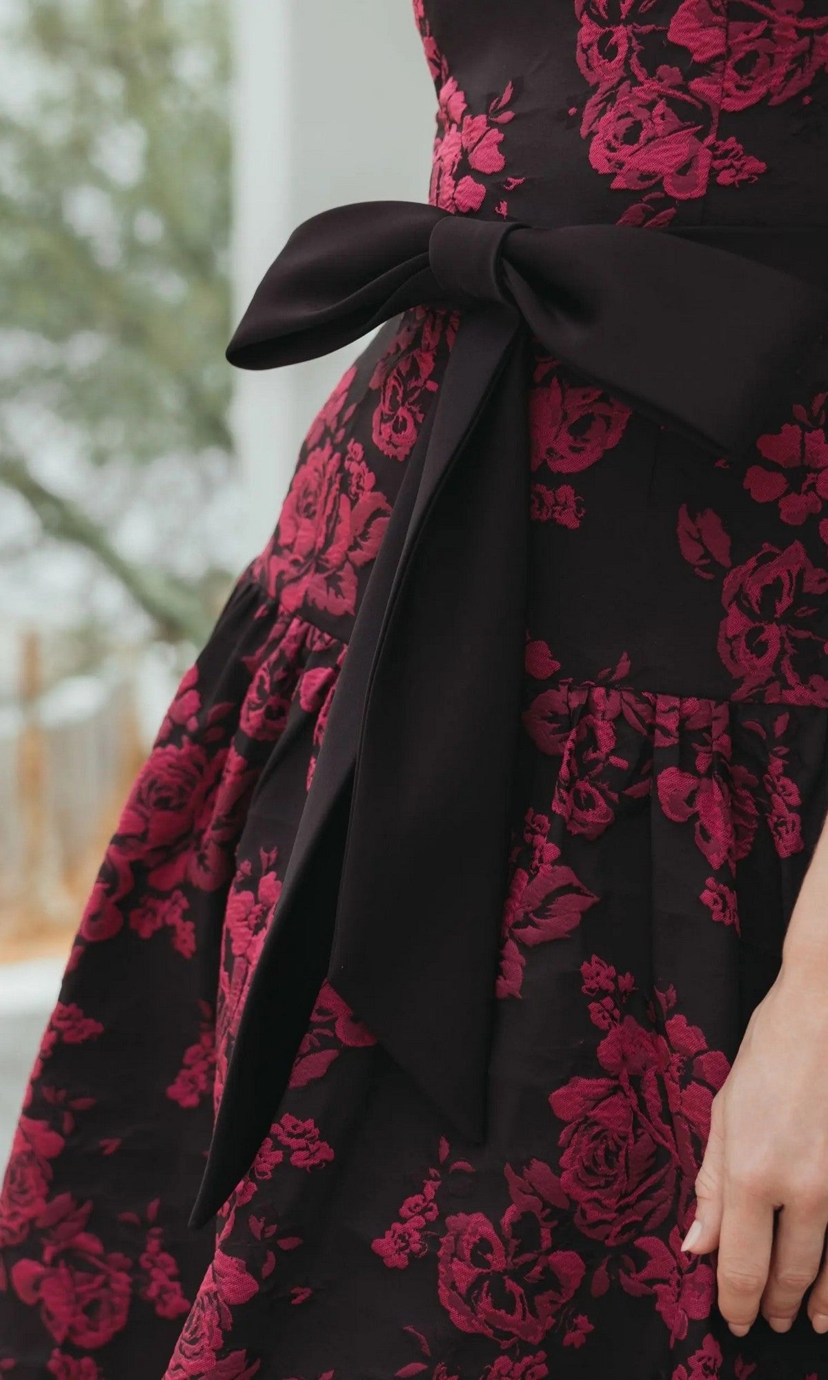 A person wears the Mac Duggal 37091 Floral Drop-Waist Party Dress, featuring a dark fabric with a red floral pattern and a large black bow at the waist.